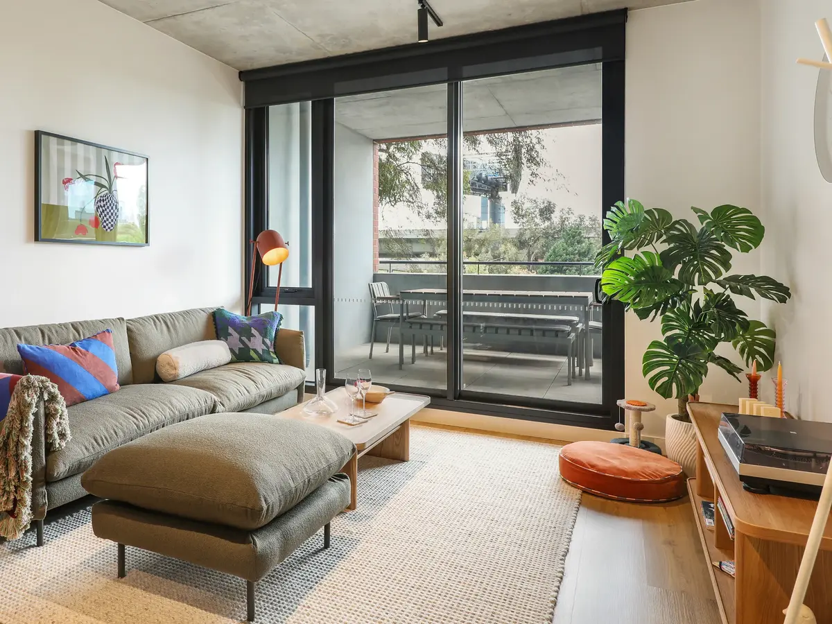 Modern living room with green sofa, colorful pillows, a floor lamp, large window opening to a balcony with outdoor furniture, and a wooden shelf with a record player and large green plant.