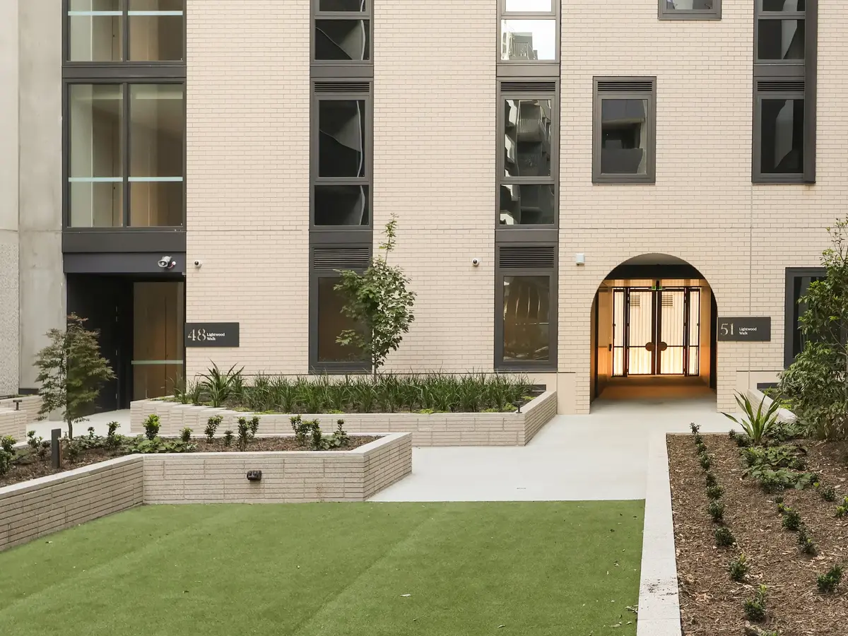 Modern beige brick apartment building entrance with landscaped garden beds and green lawn.