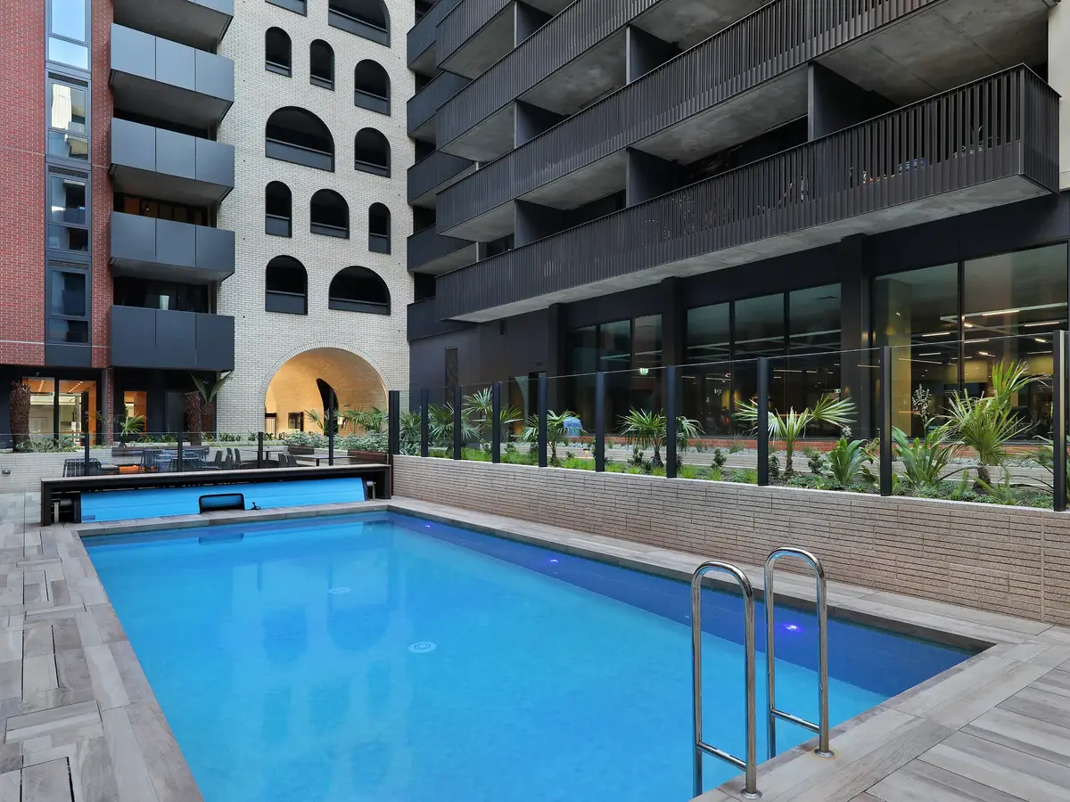 Modern outdoor swimming pool surrounded by urban apartment buildings with glass fences and small palm plants.