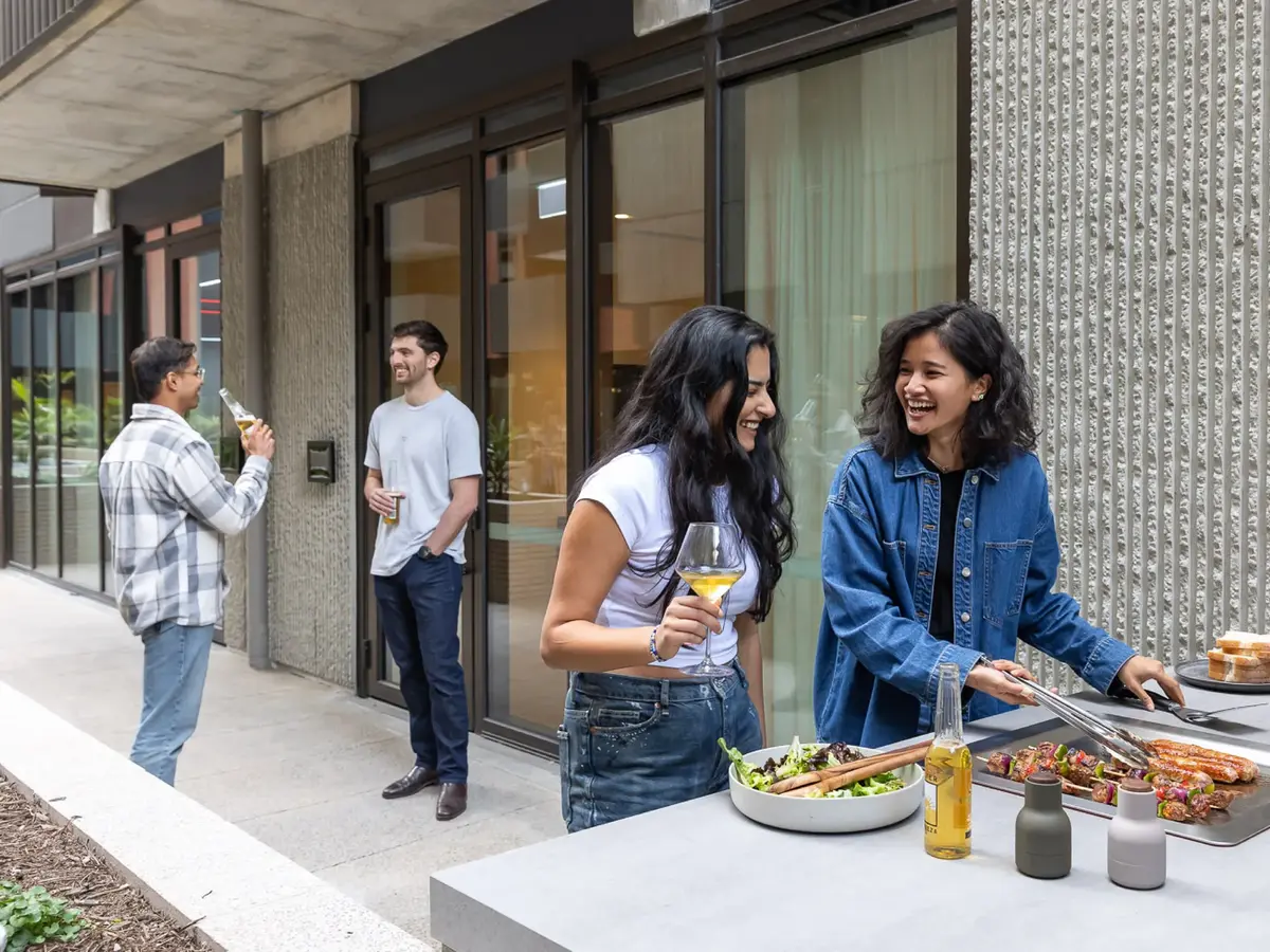 Two women smiling and preparing food at an outdoor grill while holding drinks, with two men chatting and holding bottles in the background outside a building.