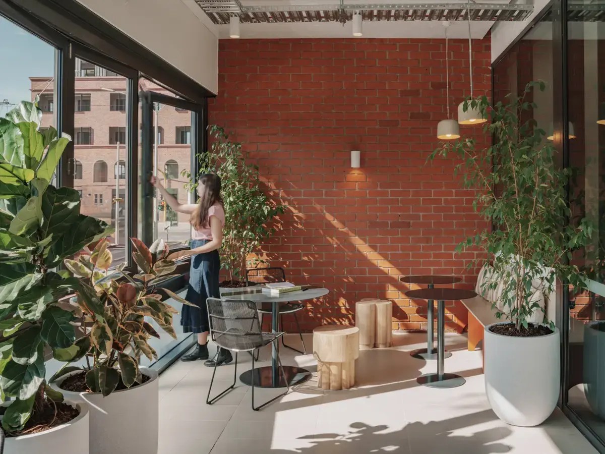 Woman opening a glass door in a modern cafe with large potted plants, brick wall, and mixed seating.