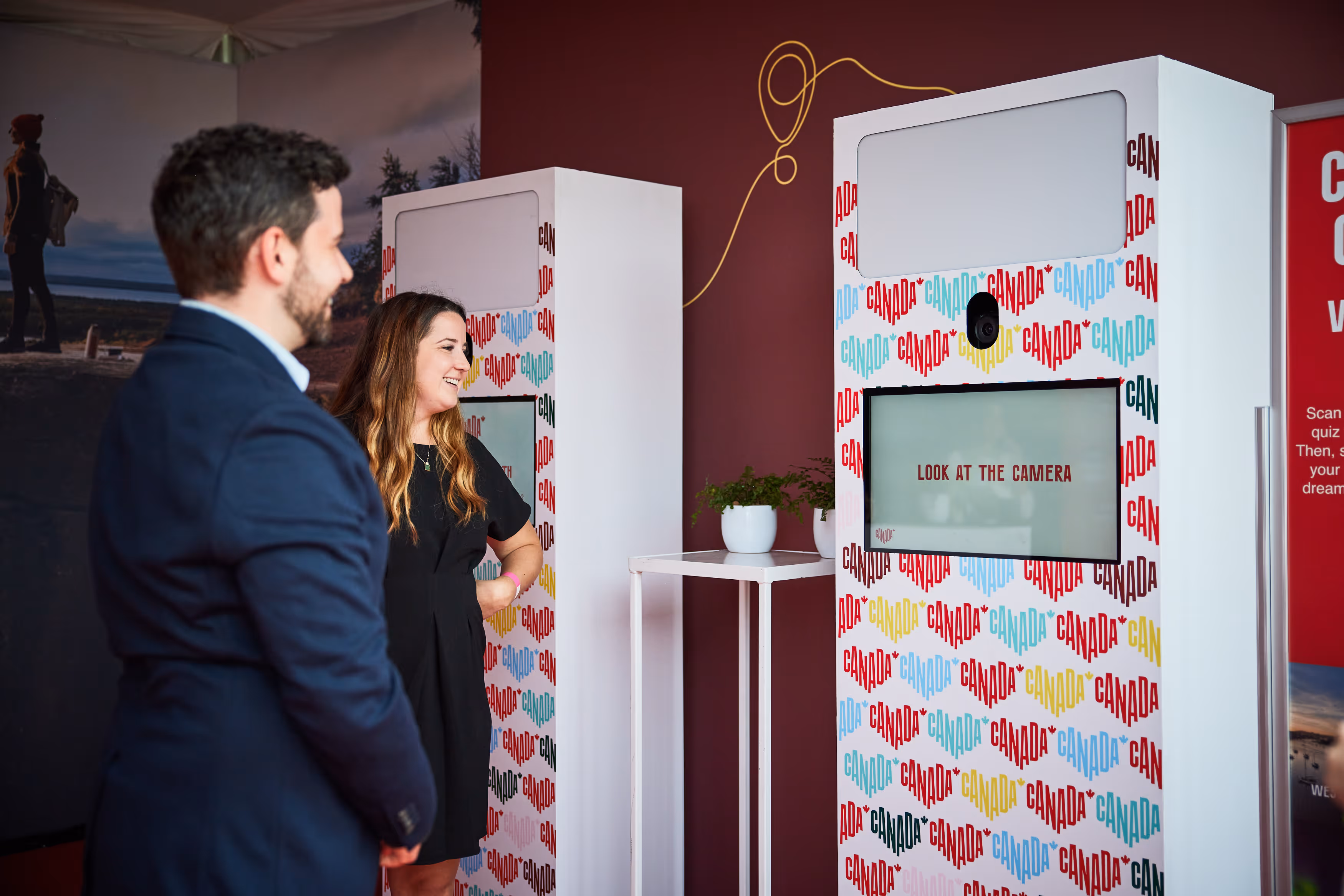 A man and woman standing smiling in front of photo booths decorated with colorful 'CANADA' text, one screen displaying 'LOOK AT THE CAMERA'.