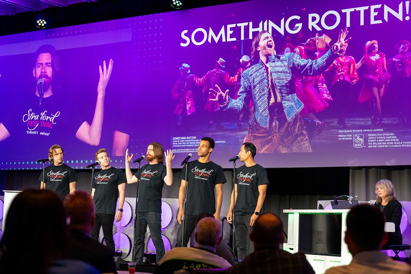 Five men wearing Stratford Festival t-shirts performing on stage with microphones, with a large screen behind them displaying an image and text for Something Rotten!