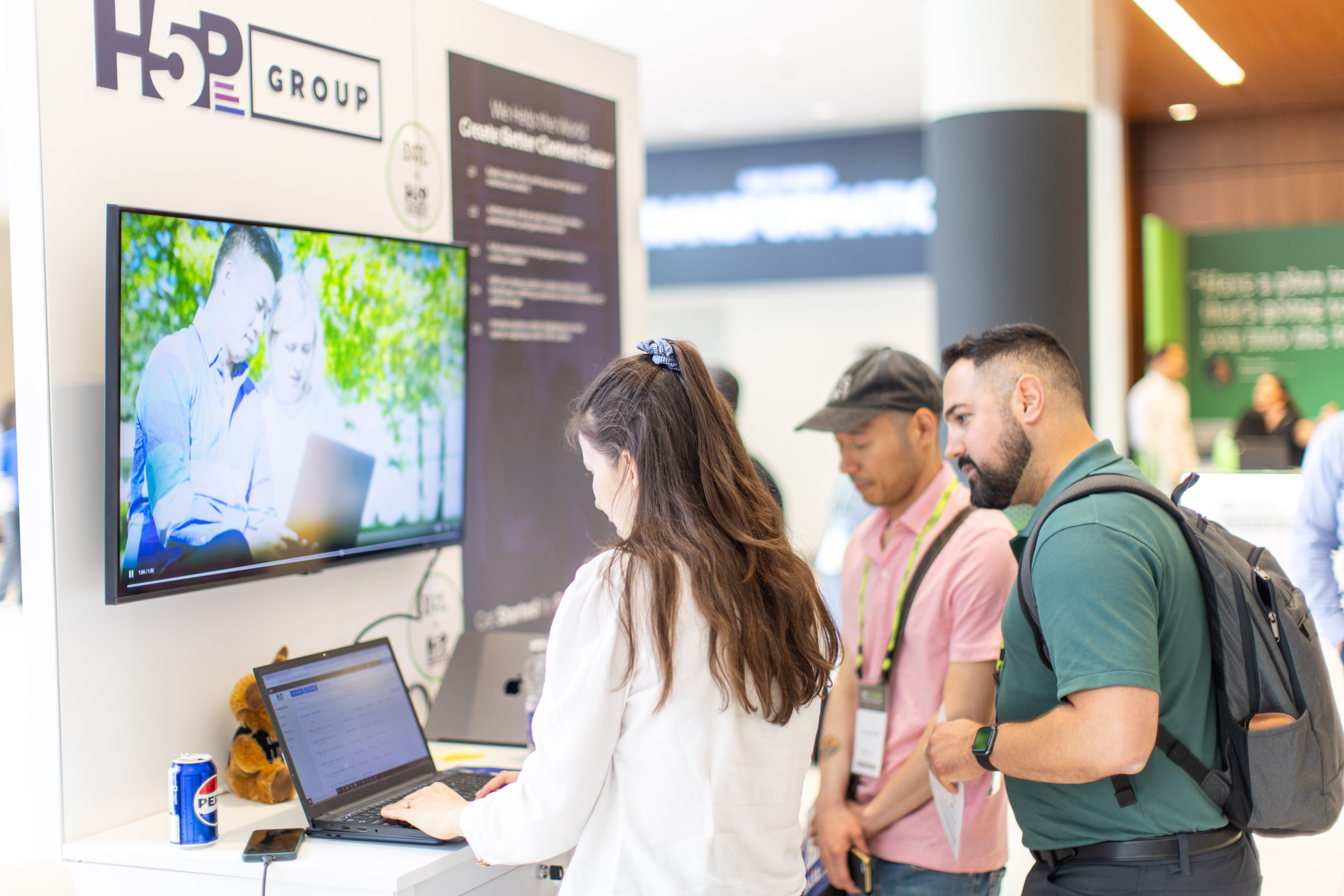 Three people interacting at a tech booth with a laptop and a monitor displaying a video of two people using a laptop outdoors.