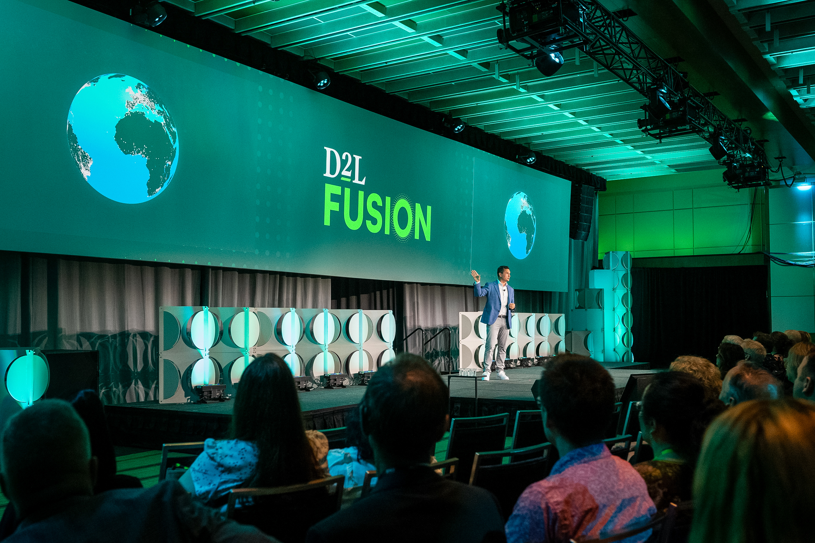 Speaker presenting on stage at D2L Fusion conference with audience watching and green-lit backdrop featuring globe images.