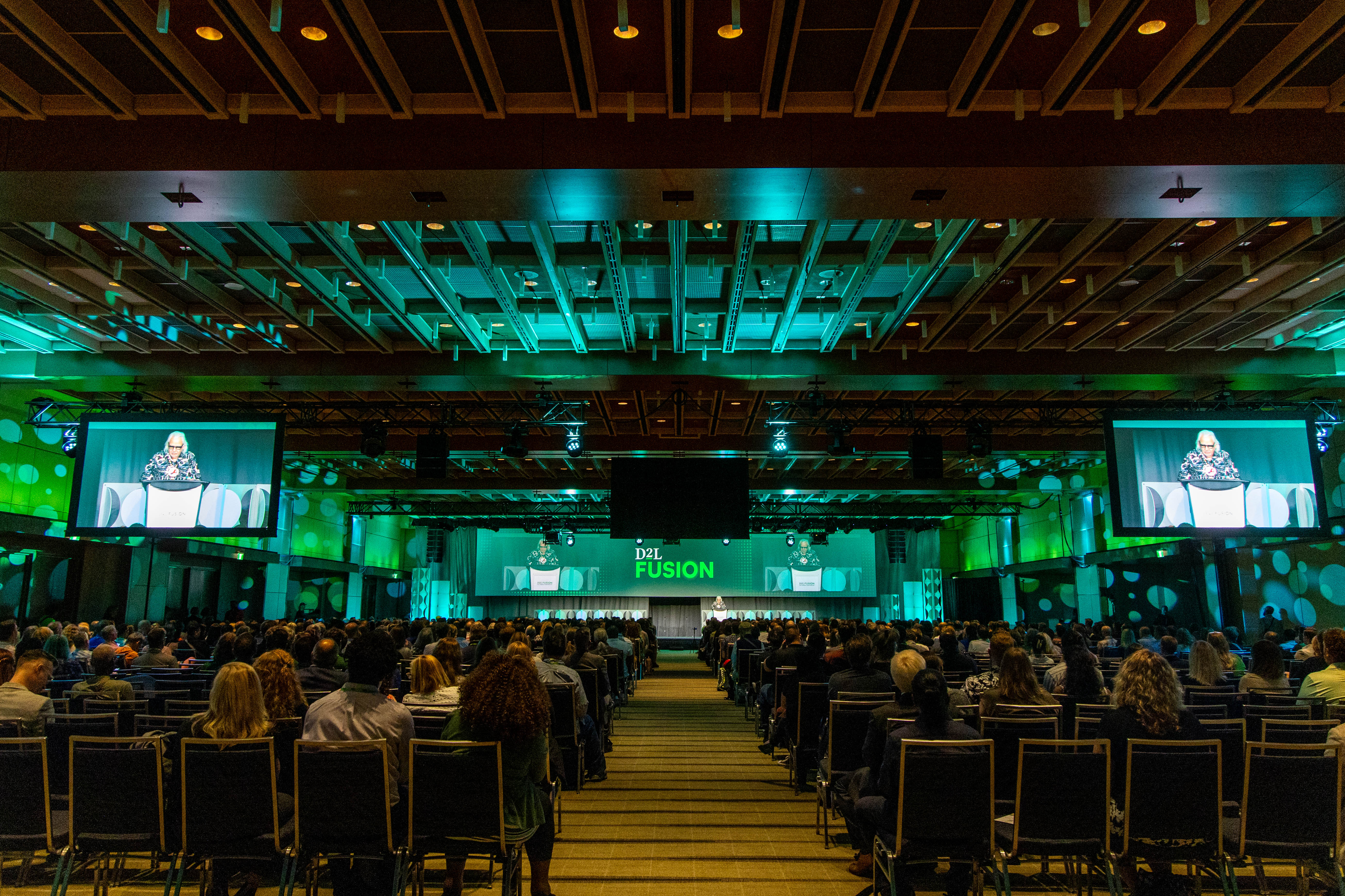 Large audience seated in a conference hall watching a speaker on stage at D2L Fusion event, with green lighting and two large screens displaying the speaker.