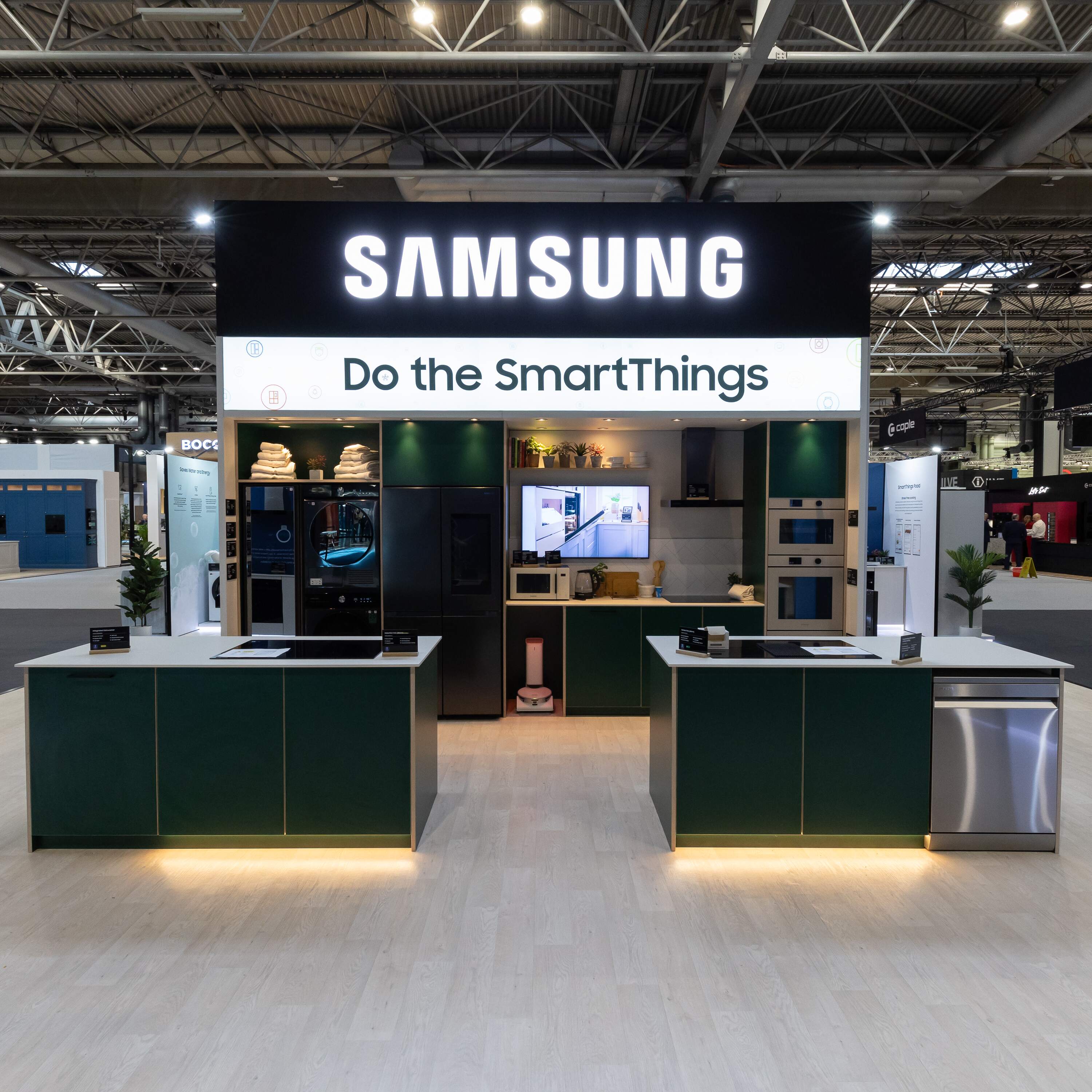 Samsung kitchen exhibit with dark green cabinets, two islands, and various smart home appliances under a sign reading 'Do the SmartThings.'