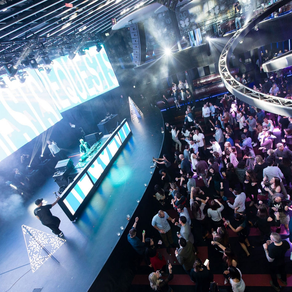 DJ performing on stage with bright light displays and a crowd dancing in a modern nightclub.