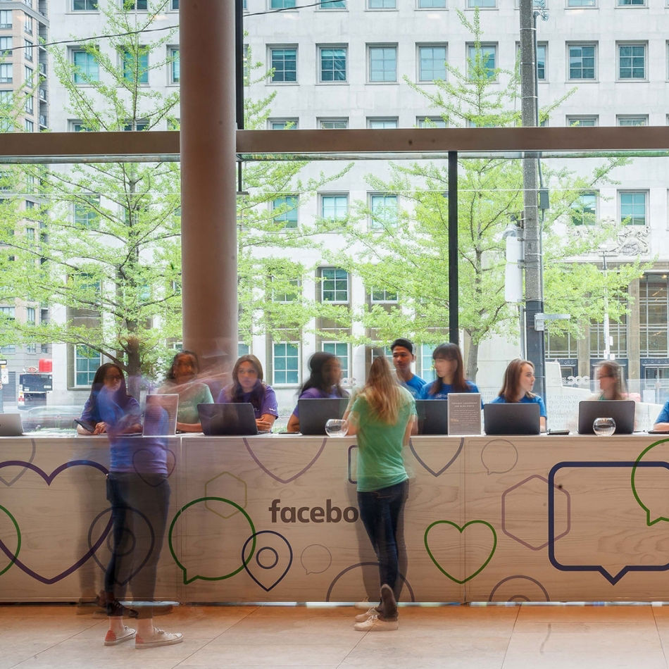 People at a brightly lit Facebook registration desk with laptops and large windows showing a cityscape.