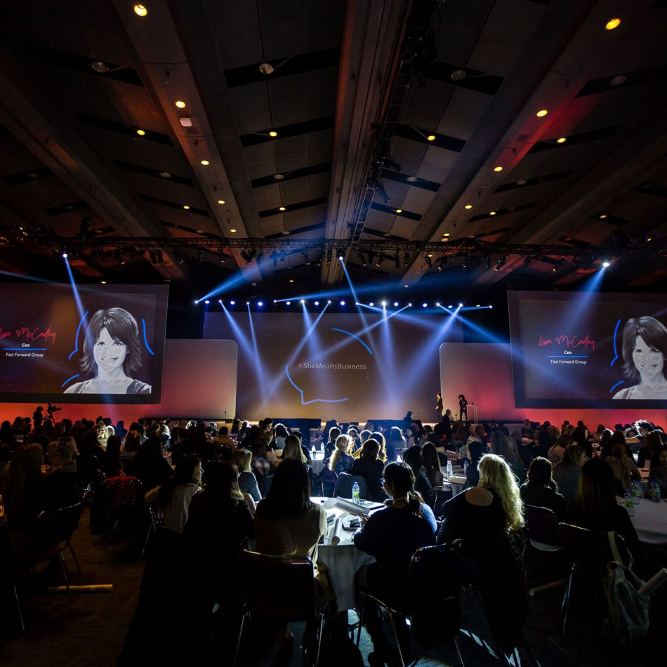 An audience seated at round tables in a large conference hall watching a presentation with two screens showing Lisa McCarthy, CEO of Fast Forward Group, under the hashtag #SheMeansBusiness.