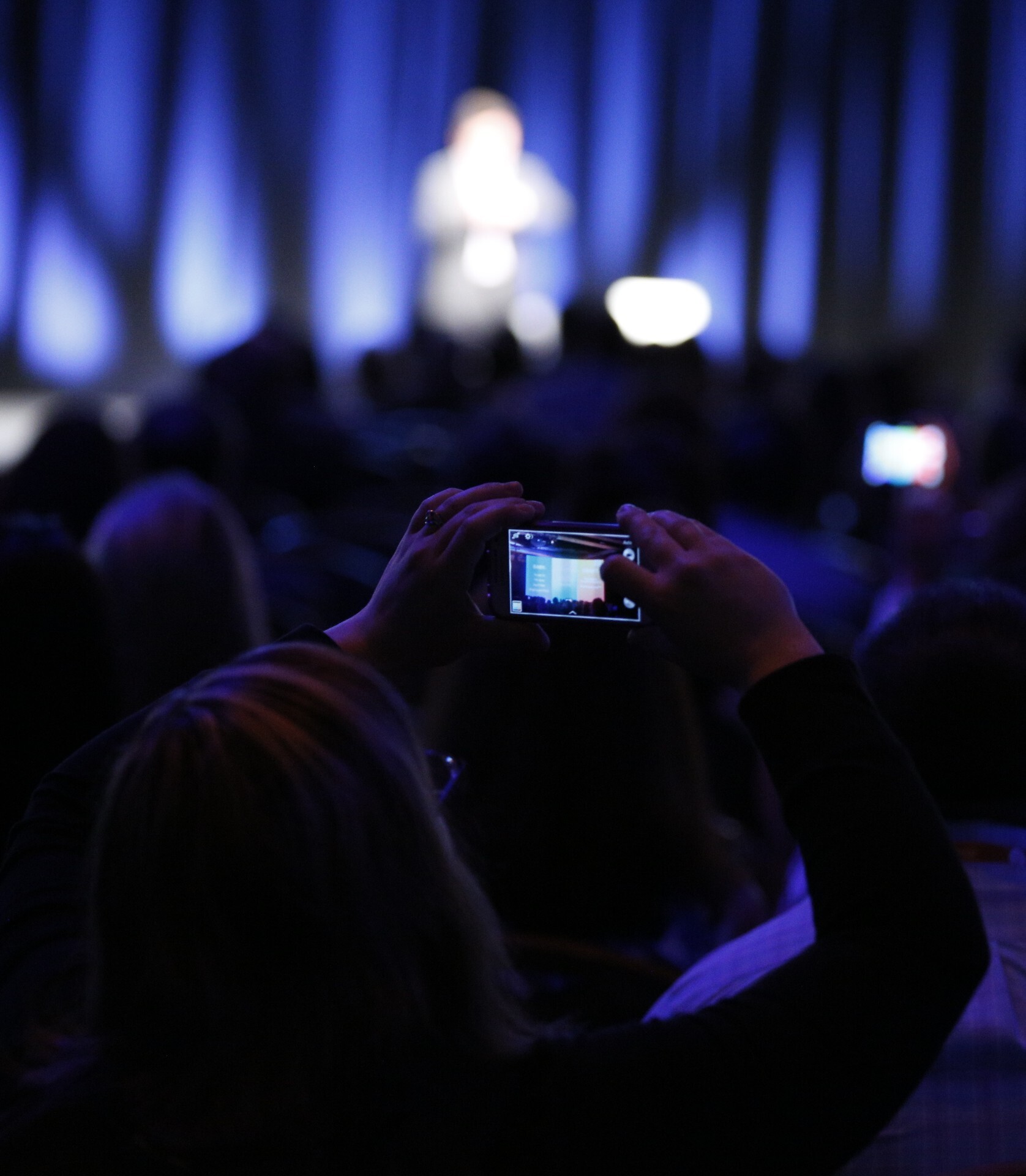 Audience member holding a phone to capture a blurry person speaking on stage with blue curtains in the background.