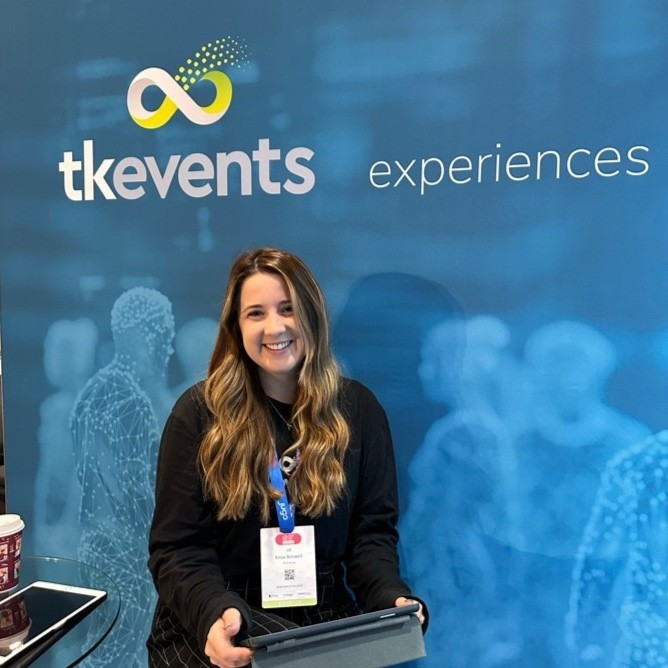 Smiling woman with long wavy hair seated in front of a blue backdrop with 'tk events experiences' logo, holding a tablet.