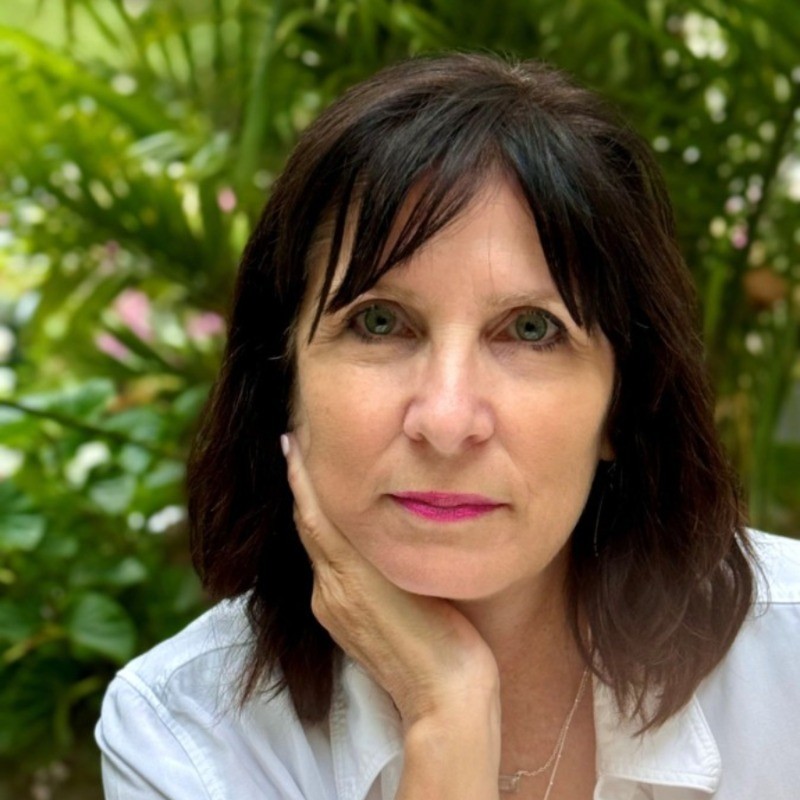 Woman with dark hair resting her chin on her hand, wearing a white shirt with blurred green foliage background.