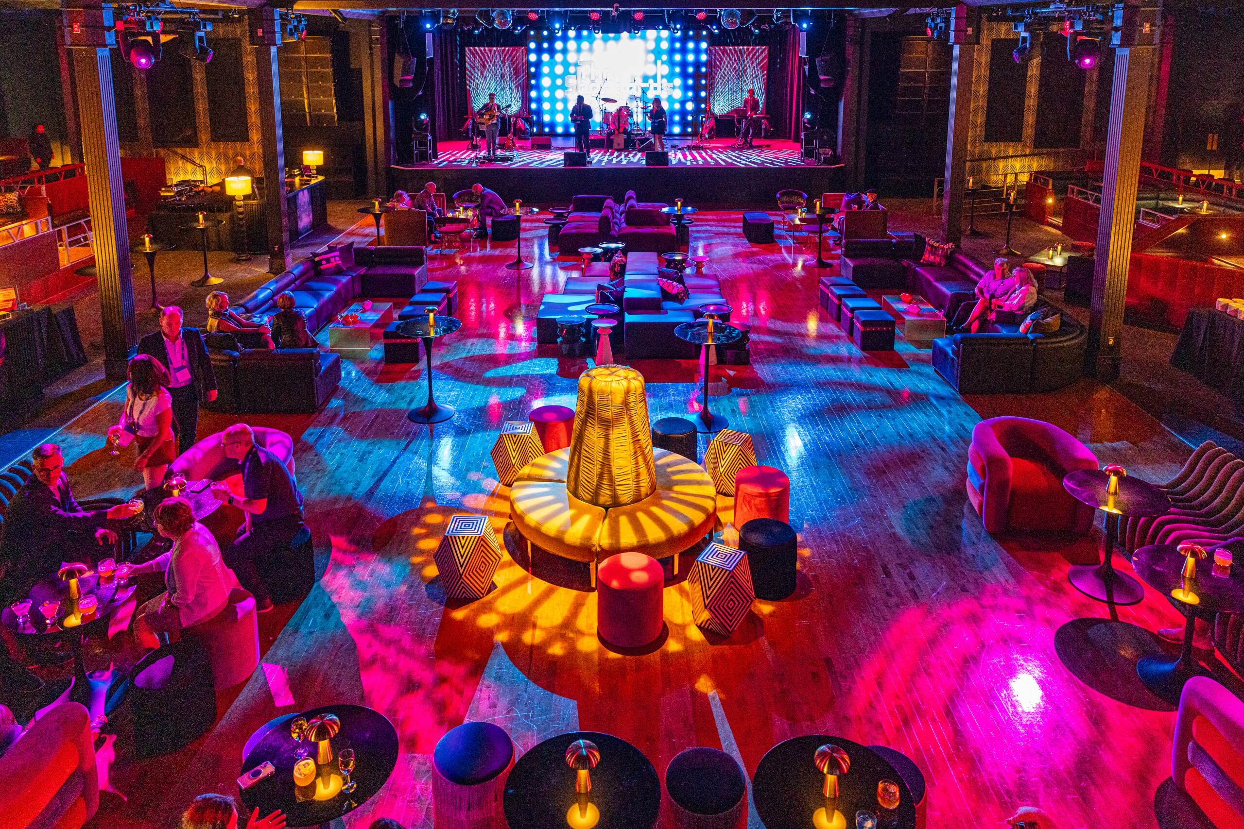 Colorfully lit lounge with people seated at tables and a band performing on a stage in the background.