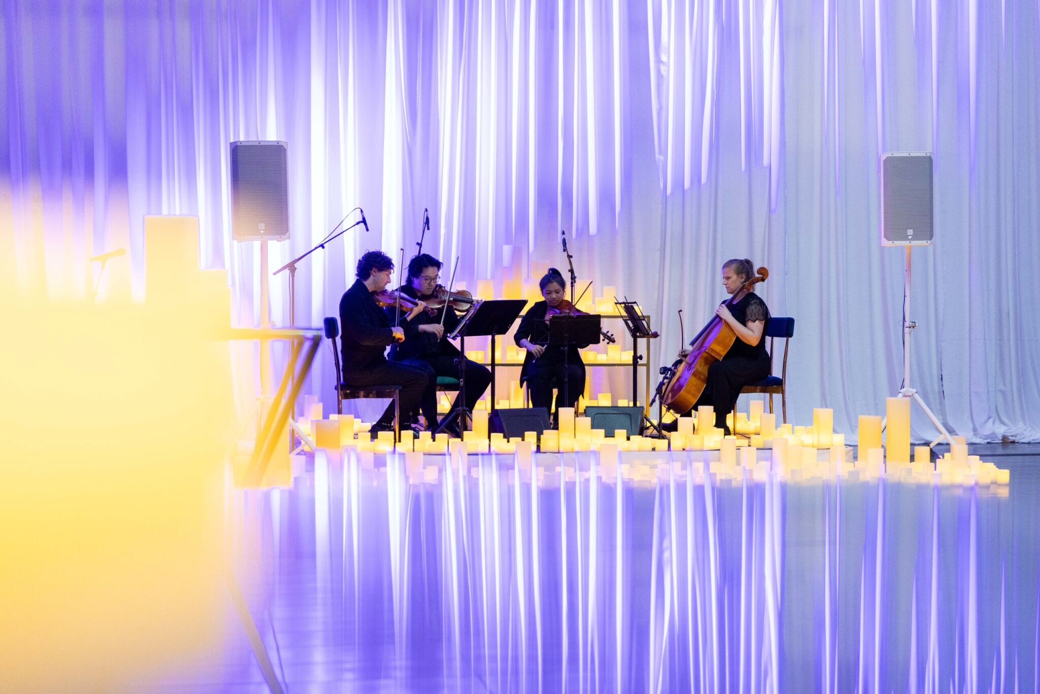 A string quartet with two violinists, one violist, and a cellist performing surrounded by numerous glowing candles and illuminated curtains.