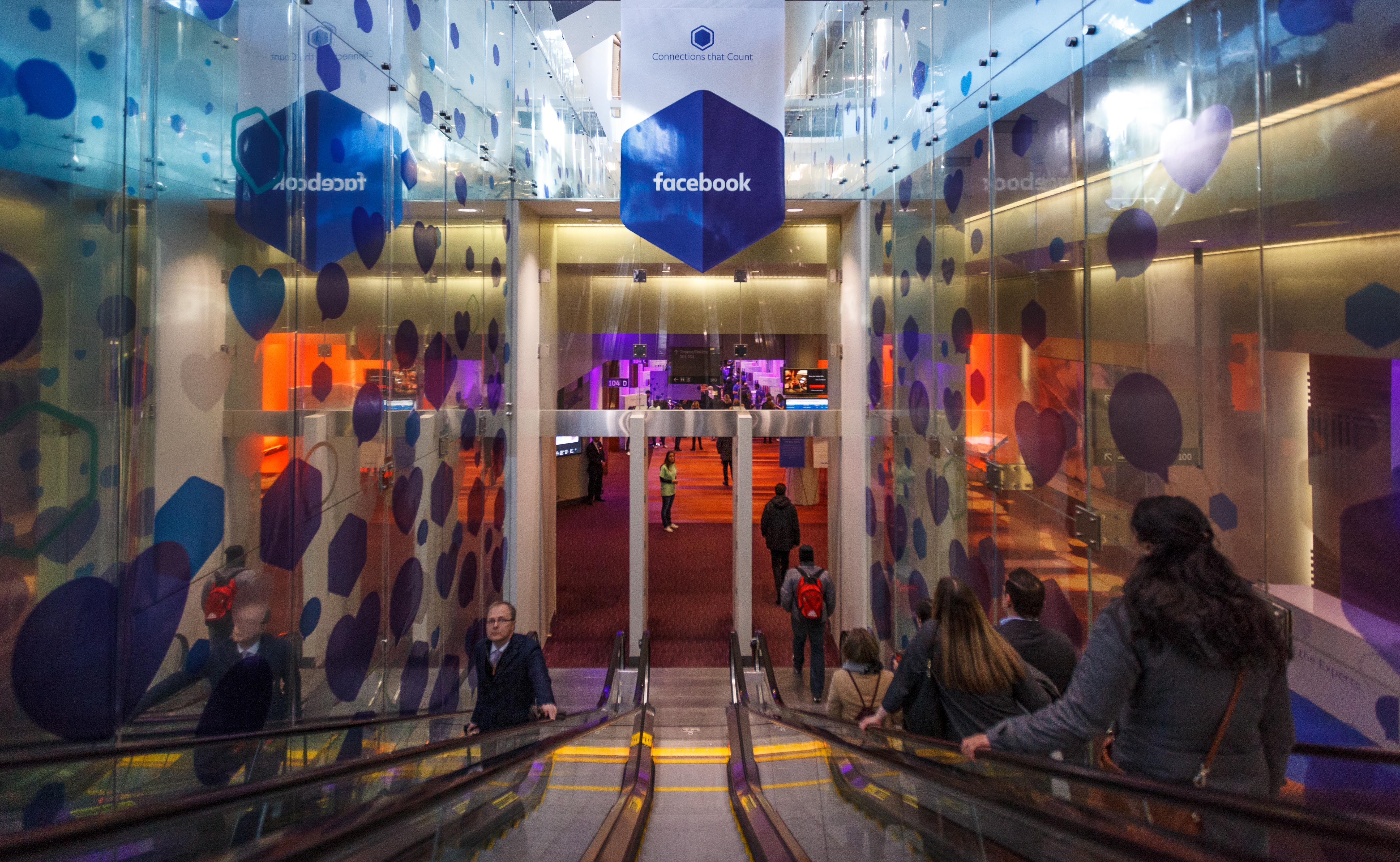People riding escalators down toward the entrance of a Facebook event venue decorated with blue and purple heart and hexagon shapes.