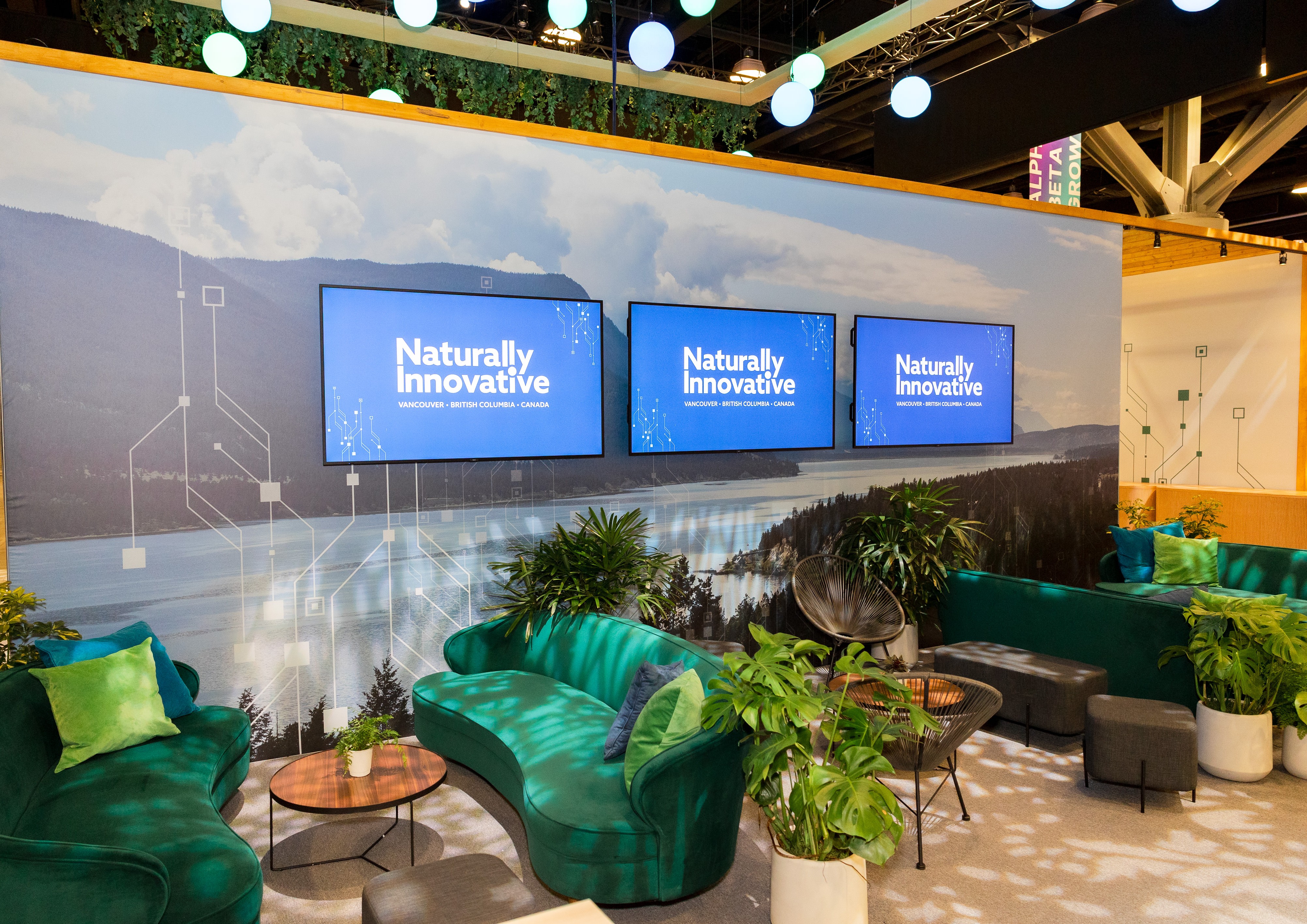 Modern lounge area with green sofas, plants, and three screens displaying 'Naturally Innovative Vancouver British Columbia Canada' against a large scenic nature backdrop.