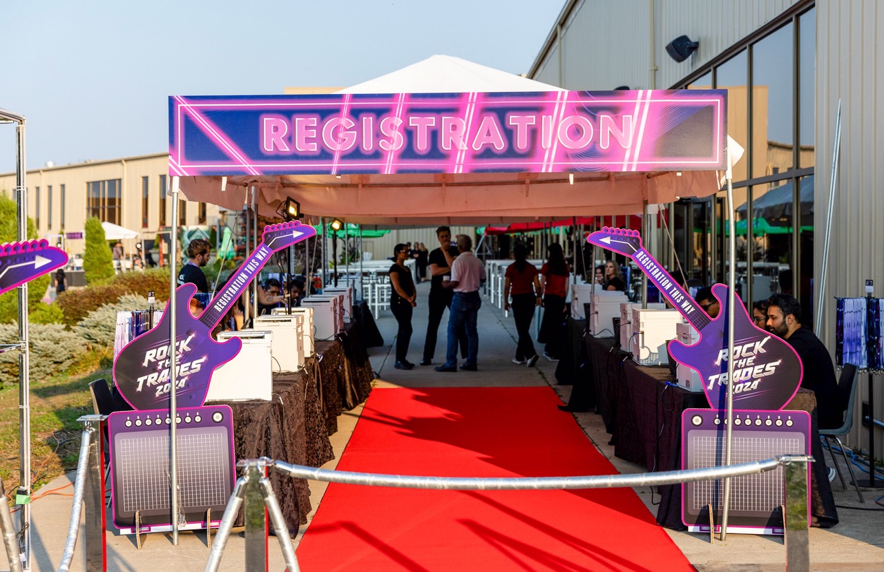 Outdoor event registration area with a red carpet, neon 'Registration' sign, and guitar-shaped signs reading 'Rock the Trades 2024'.