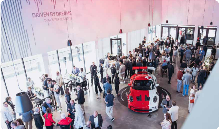 Crowd of people socializing in a spacious showroom with large windows and a red and white race car displayed in the center.