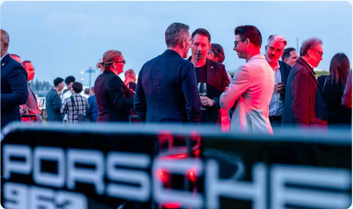 Group of people socializing outdoors in evening light near a Porsche 962 car.