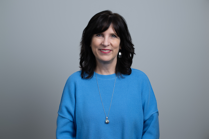 Smiling woman with shoulder-length dark hair wearing a blue sweater and silver jewelry against a gray background.