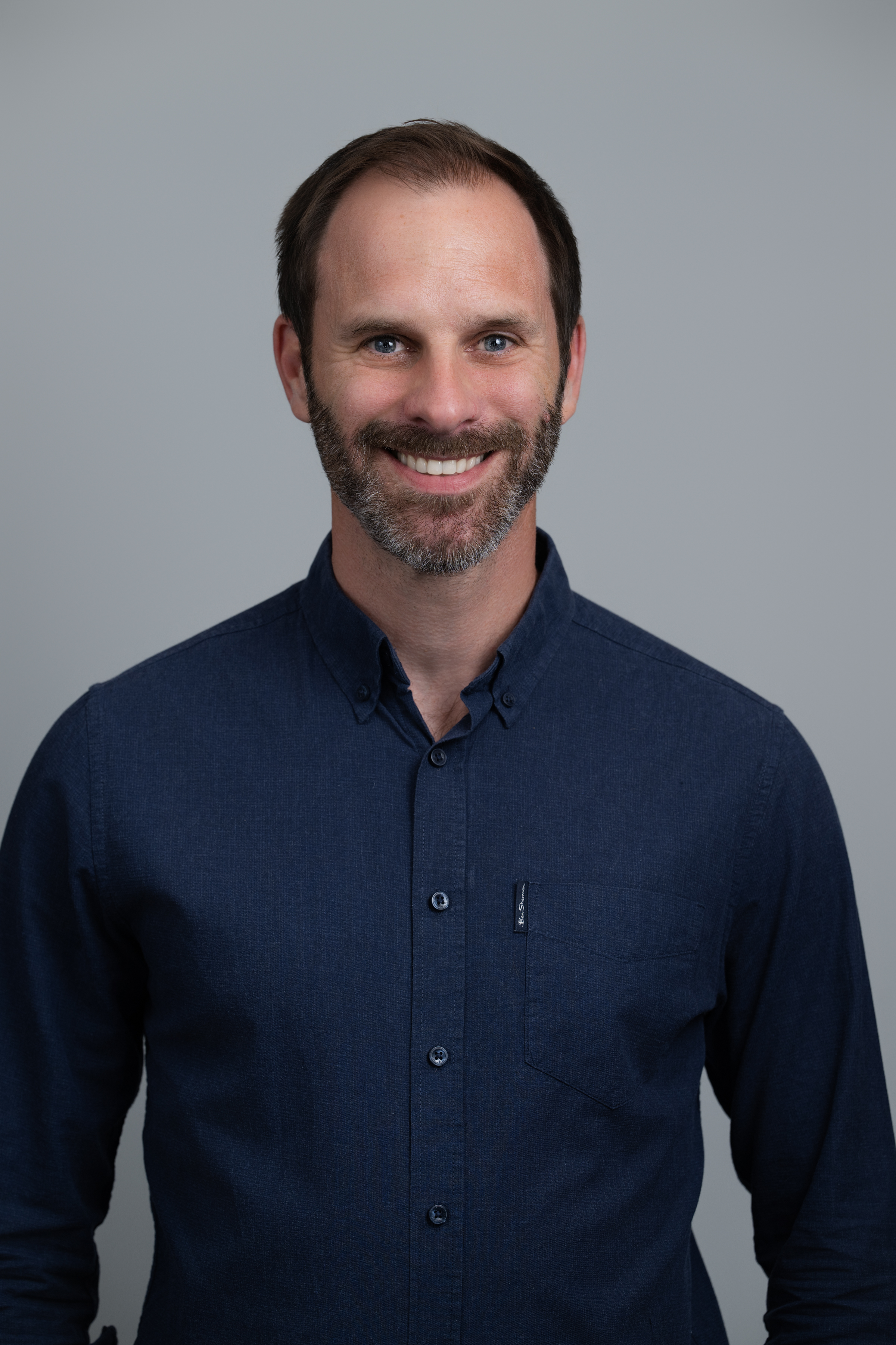 Smiling man with short brown hair and beard wearing a dark blue button-up shirt against a plain gray background.