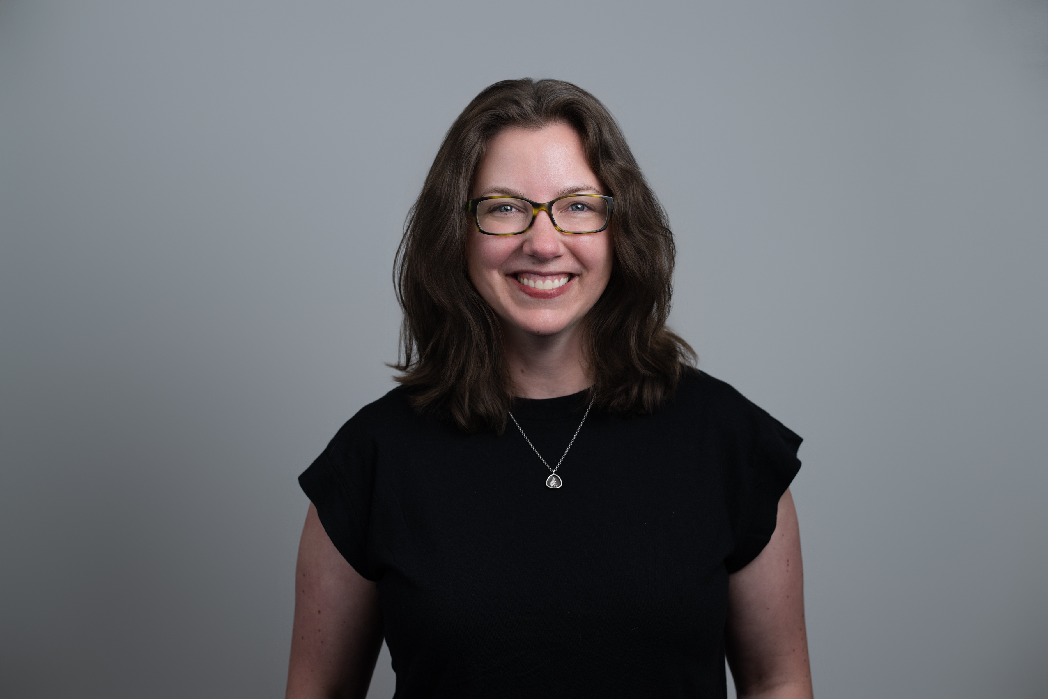 Smiling woman with shoulder-length brown hair, wearing glasses, a black shirt, and a pendant necklace against a gray background.