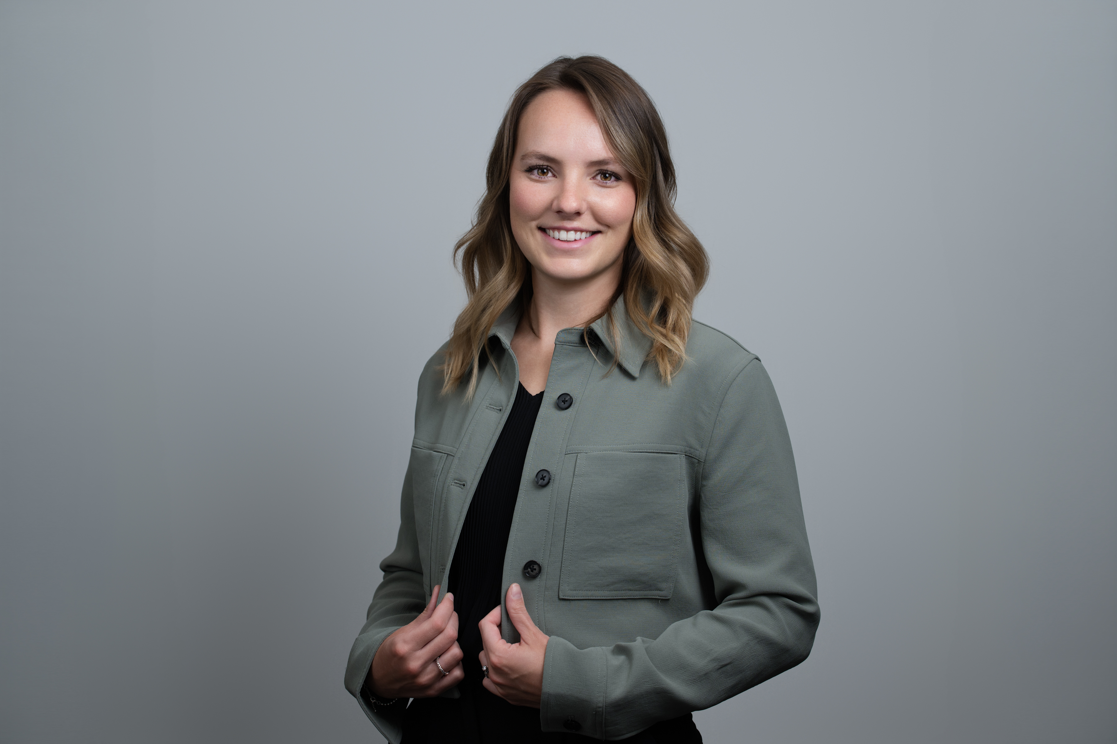 Smiling woman with shoulder-length wavy hair wearing a green buttoned jacket over a black top, standing against a gray background.