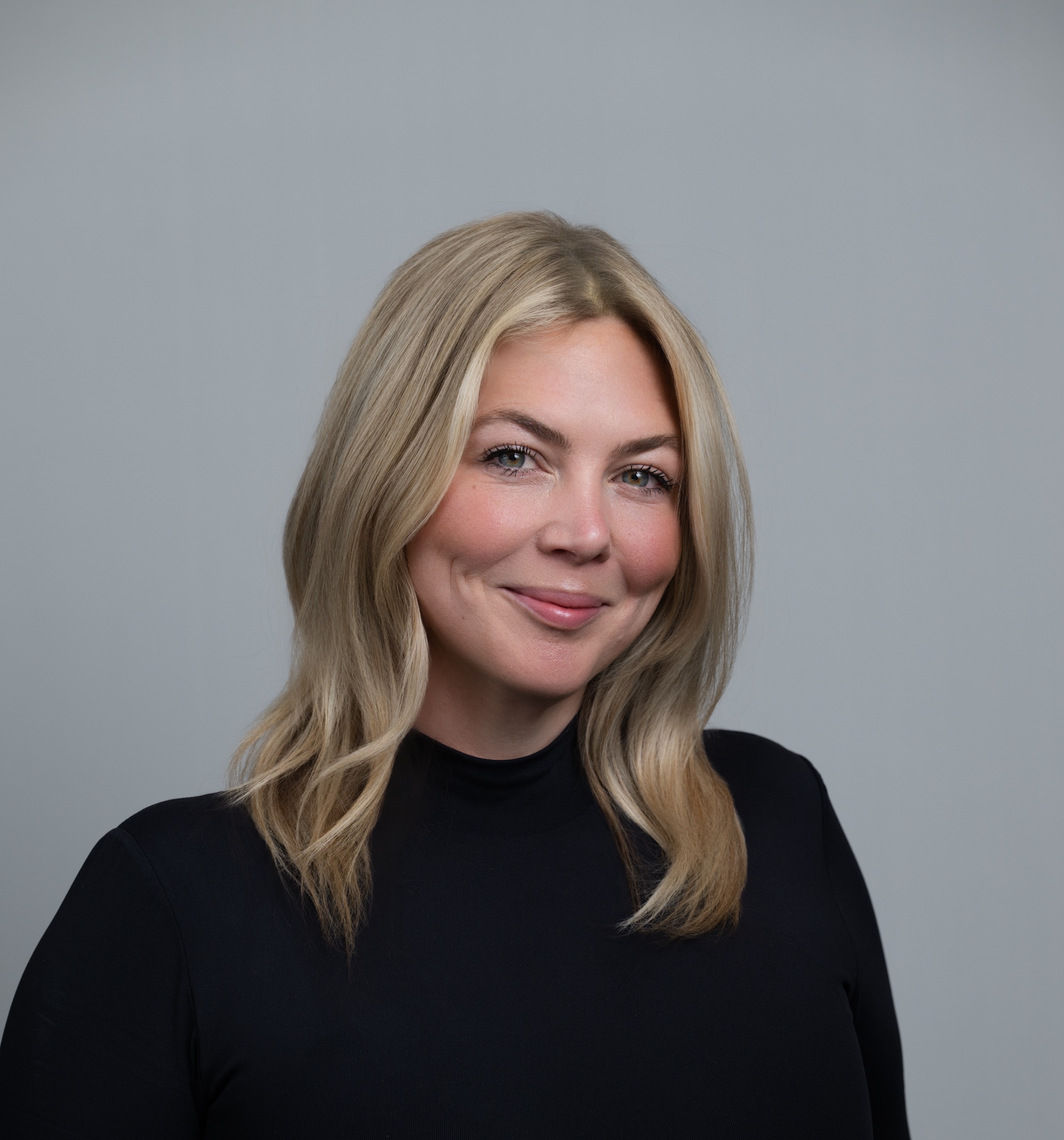 Smiling woman with shoulder-length blonde hair wearing a black top against a gray background.