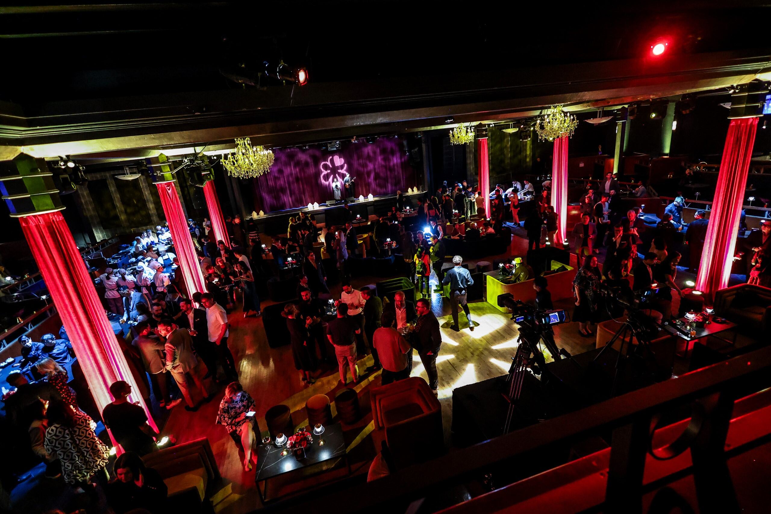 Crowd of people socializing in a dimly lit event venue with red-lit pillars and chandeliers.