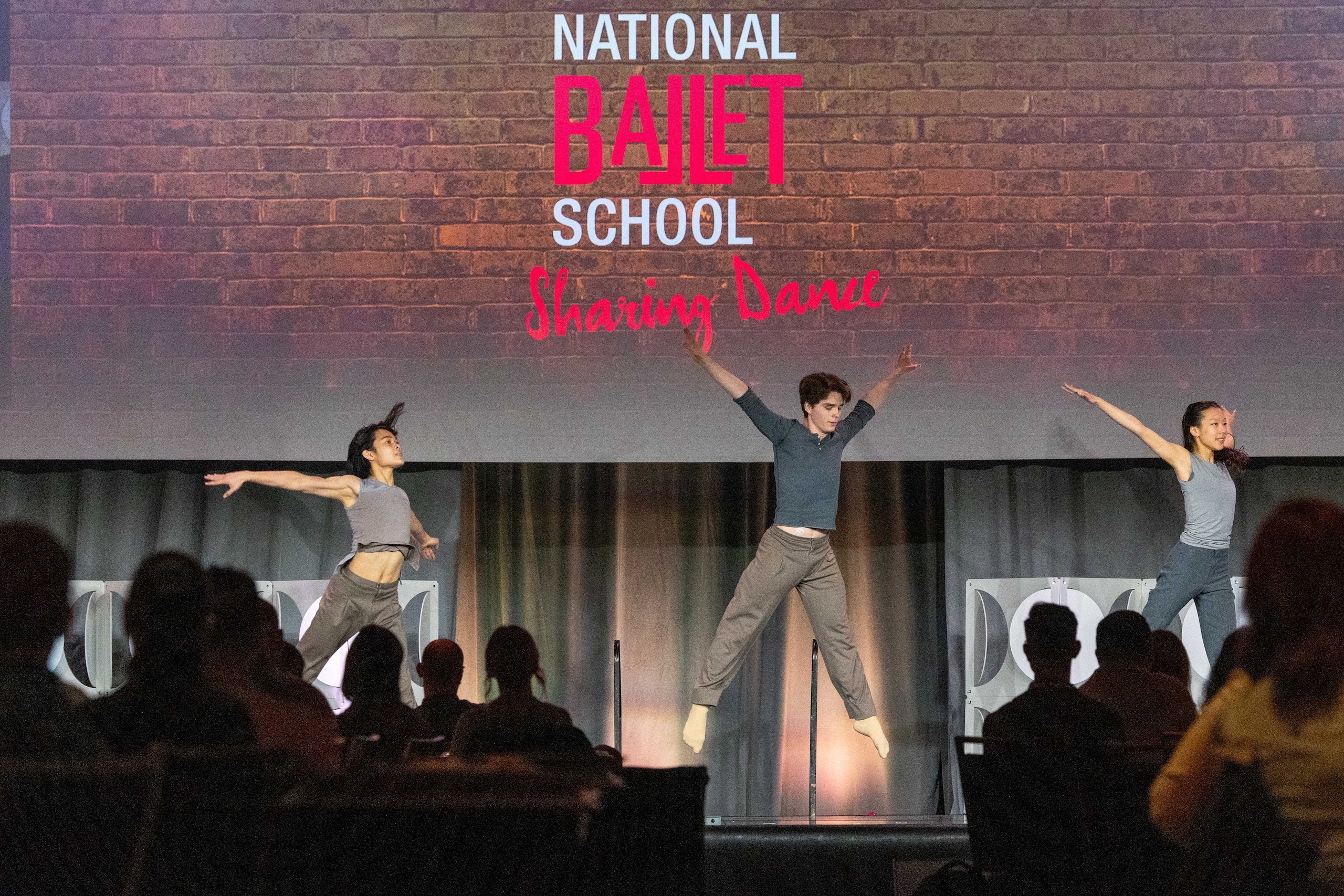 Three ballet dancers performing a jump on stage with a backdrop reading 'National Ballet School Sharing Dance' and an audience watching.