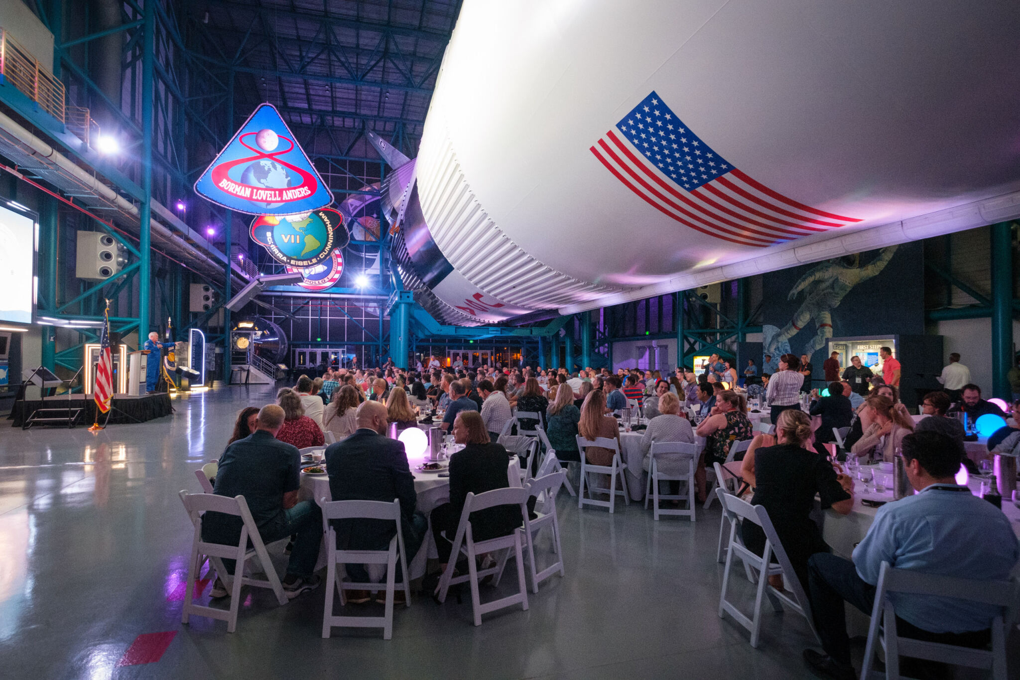 People seated at round tables attend a formal event inside a space museum with a large rocket overhead and NASA mission patches hanging.