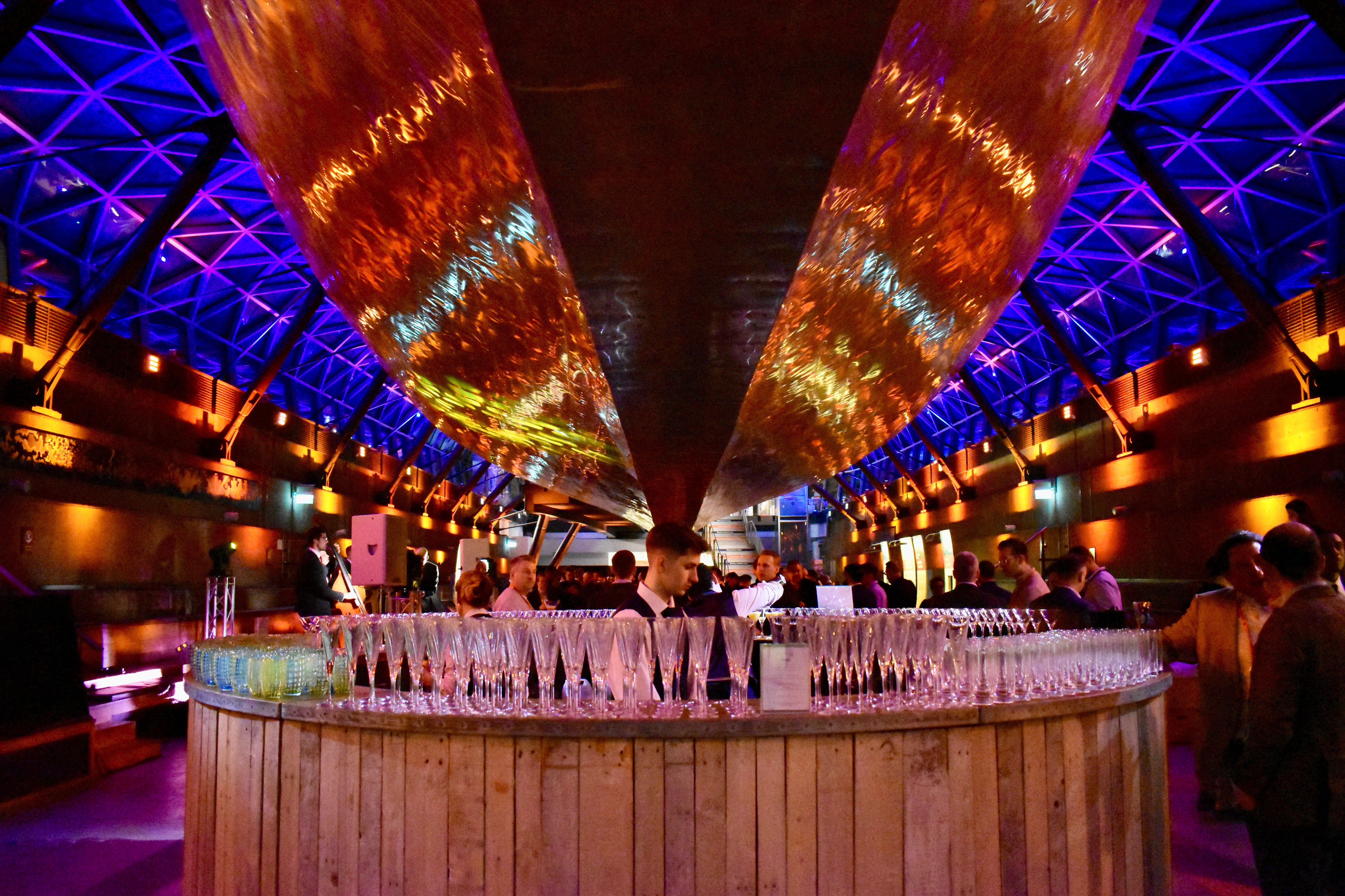 Bar setup with rows of empty champagne glasses and people mingling under a ceiling with blue and orange lighting.