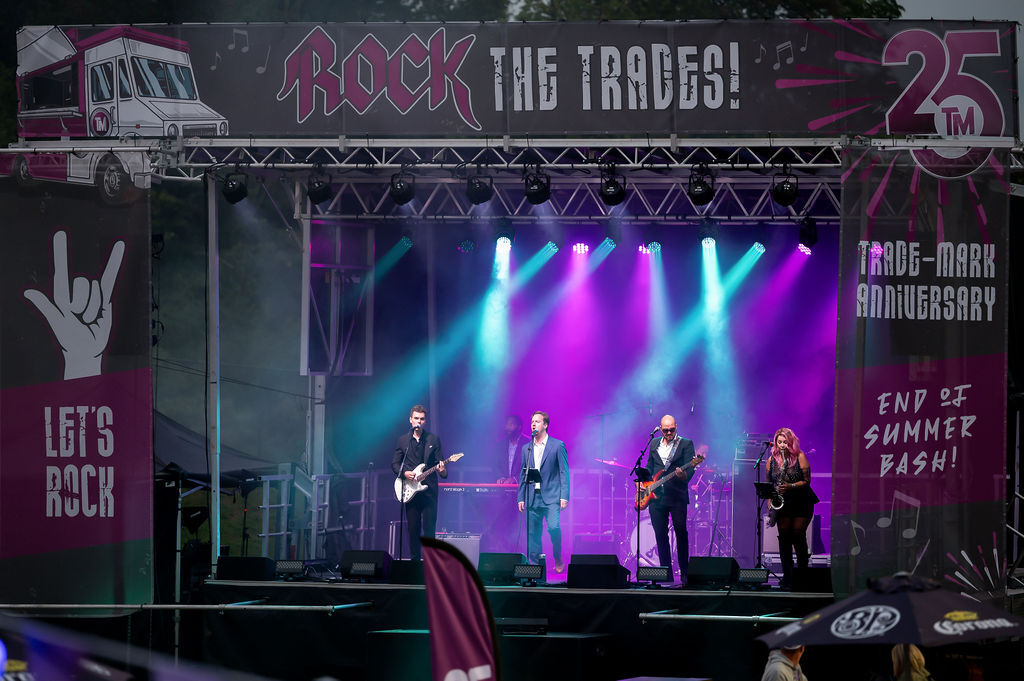Band performing on stage with colorful lights at a Rock the Trades 25th Trade-Mark Anniversary end of summer bash event.