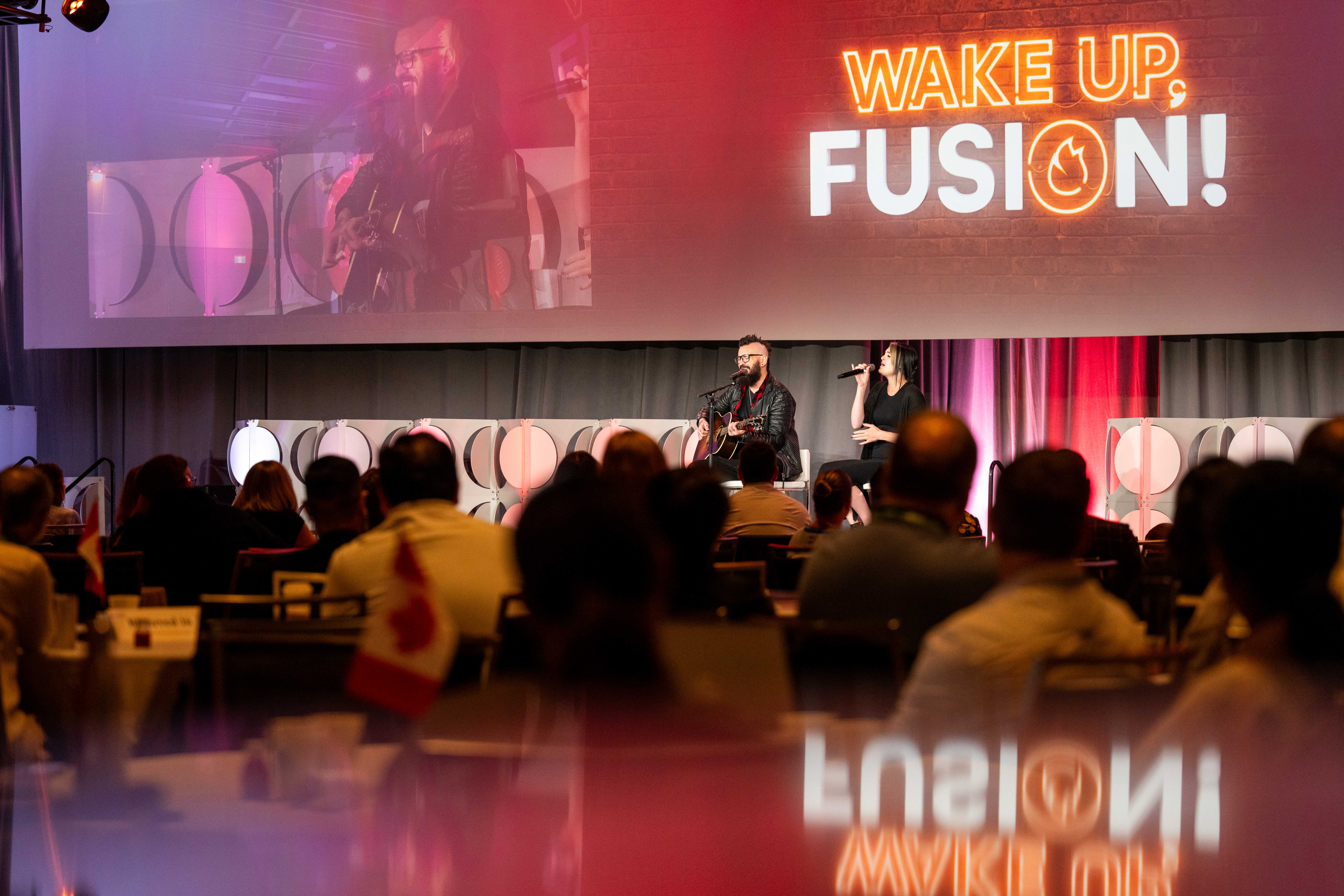A man playing guitar and a woman singing on stage in front of an audience, with a large screen displaying 'WAKE UP, FUSION!' behind them.