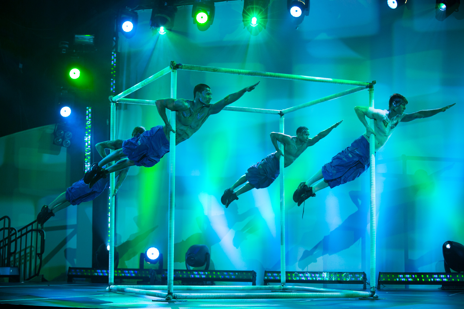 Four male acrobats performing a horizontal flag pose gripping vertical poles of a large apparatus on a blue-green lit stage.