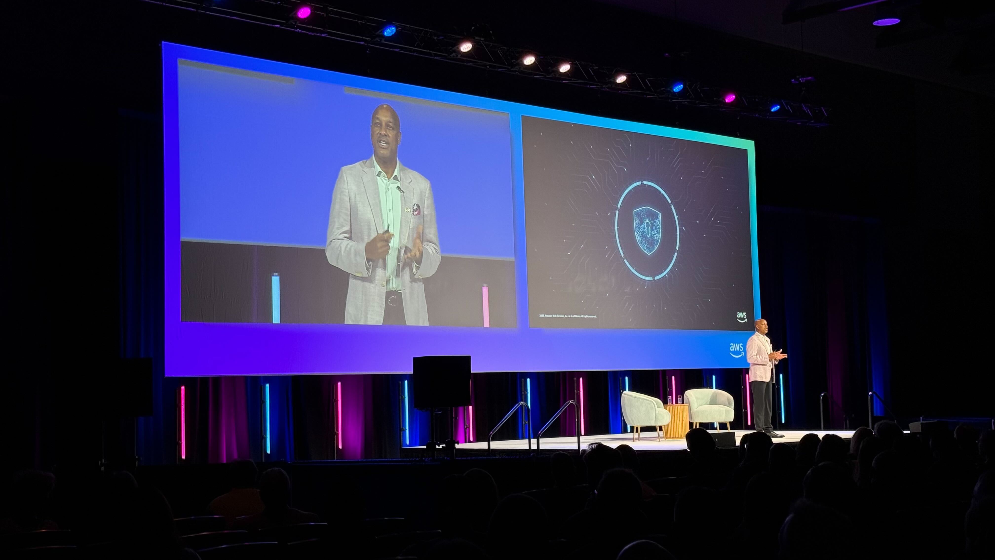 Man in a light gray blazer speaking on stage at an AWS event with a large screen behind showing his image and a digital shield symbol.