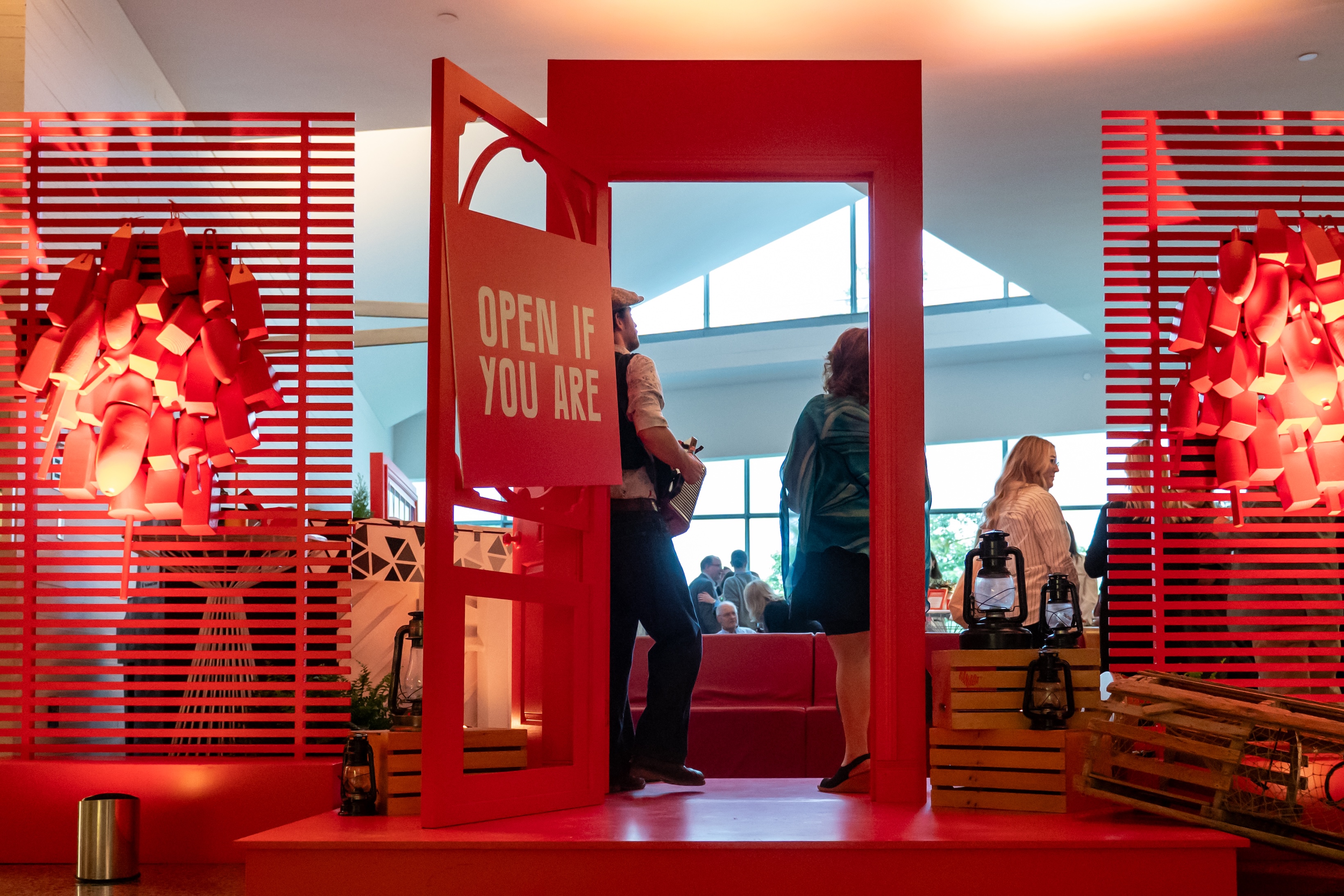 Red door with a sign saying 'OPEN IF YOU ARE' with people inside a bright room featuring large windows.
