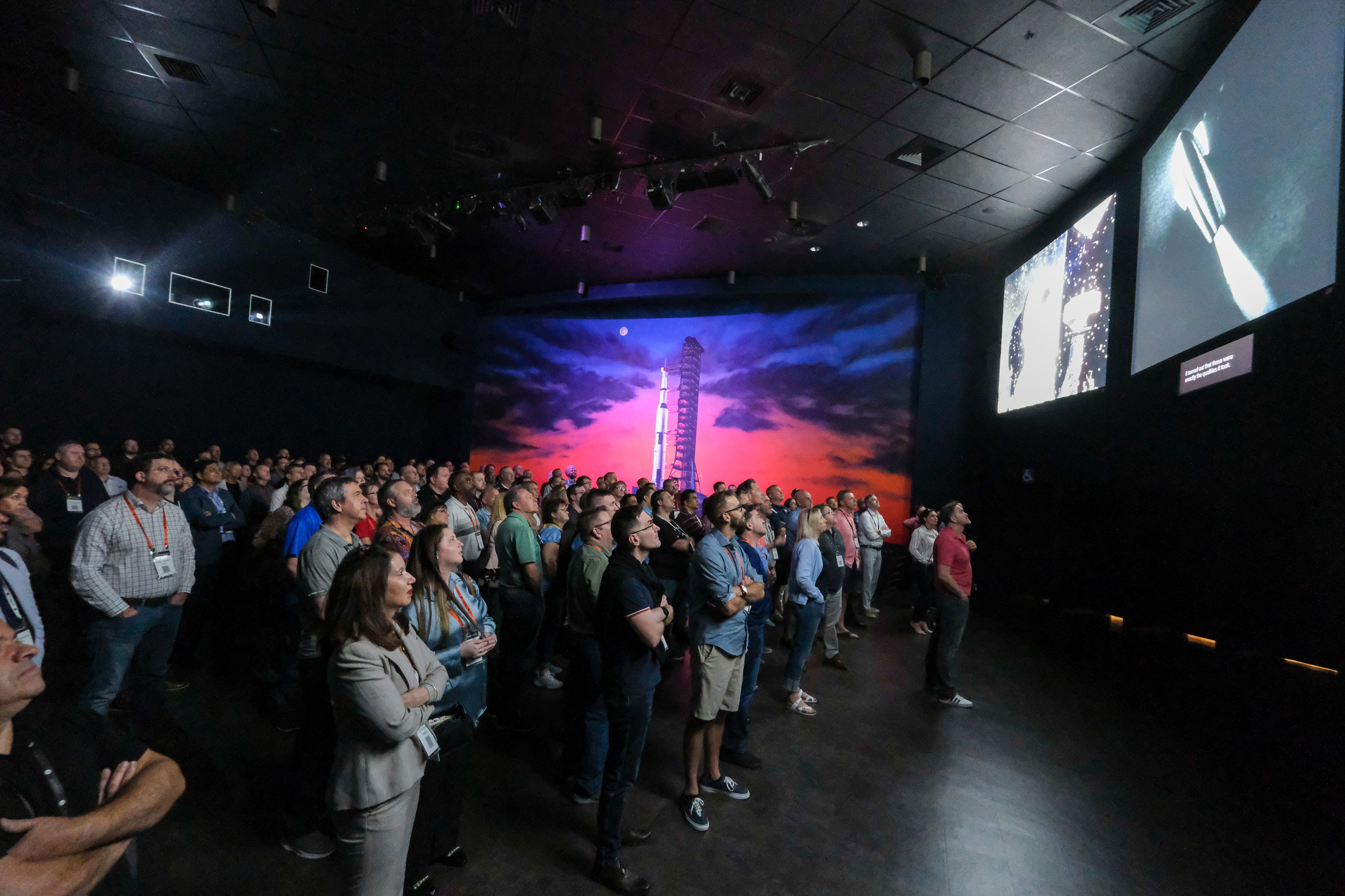 Large audience attentively watching a space launch presentation in a dimly lit room with a mural of a rocket and colorful sky on the back wall.