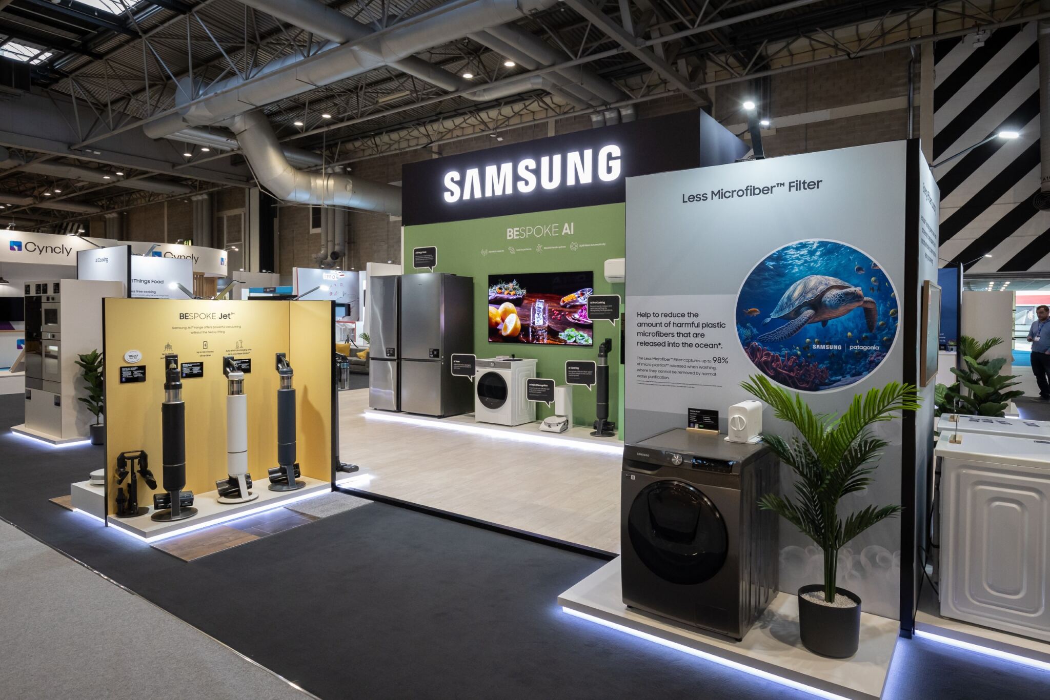 Samsung exhibition booth displaying home appliances including refrigerators, washing machines, and vacuum cleaners with a sign about less microfiber pollution featuring a sea turtle image.