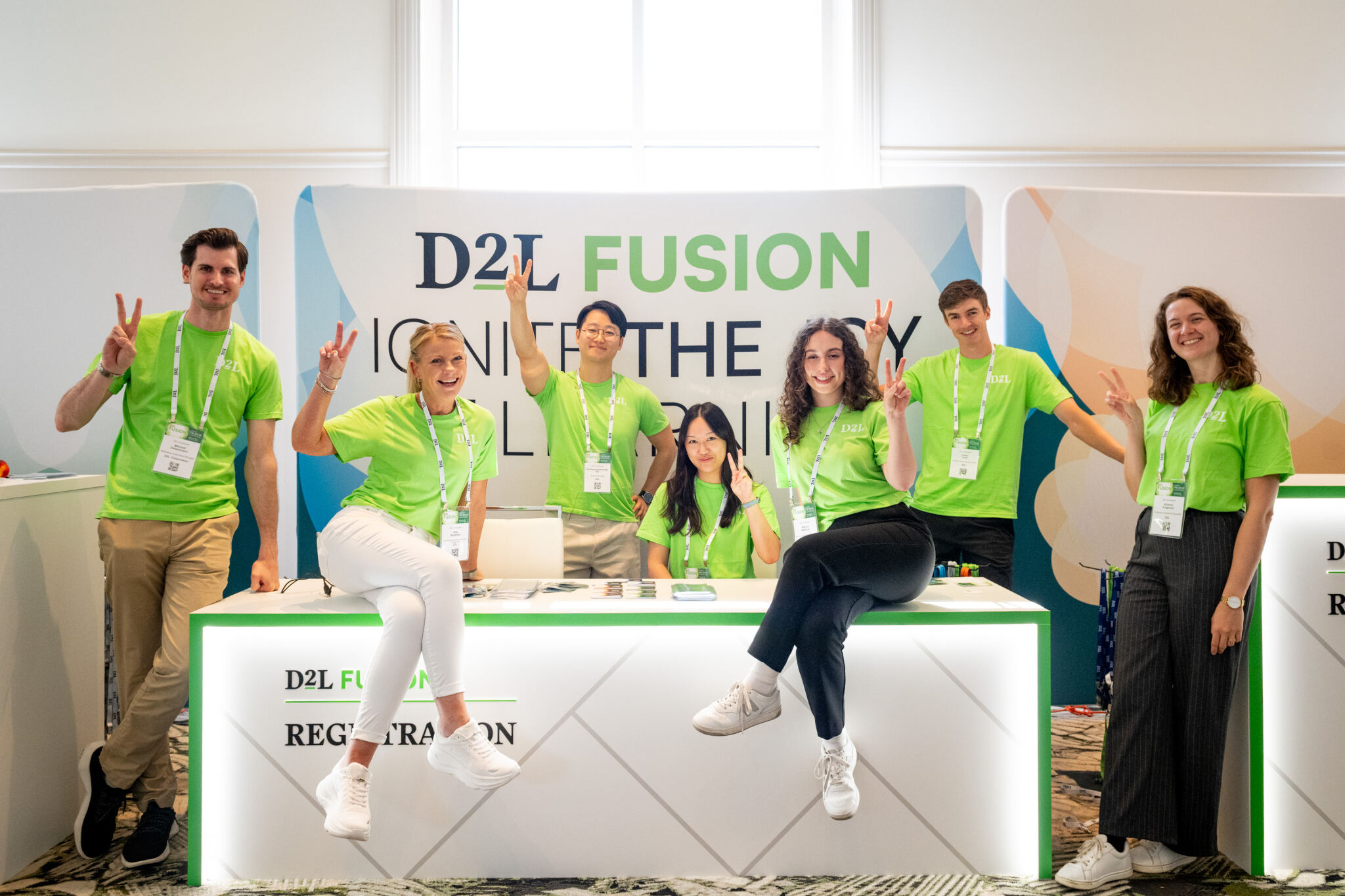 Seven smiling people wearing bright green D2L shirts posing with peace signs at a D2L Fusion registration booth.