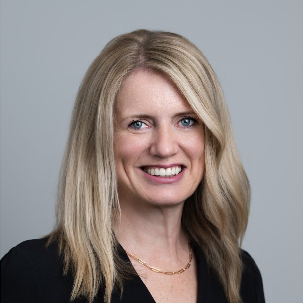 Smiling woman with long blonde hair wearing a black top and a gold chain necklace against a plain gray background.