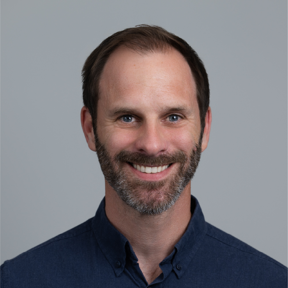 Smiling man with short brown hair, beard, and blue eyes wearing a dark blue button-up shirt against a gray background.