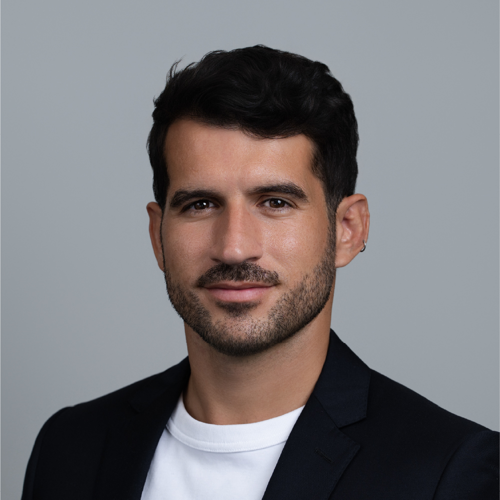 Portrait of a man with dark hair and beard wearing a black blazer and white t-shirt against a gray background.