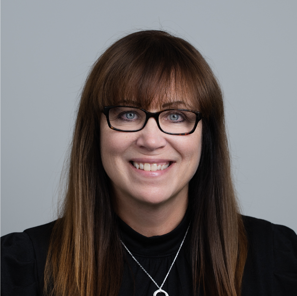 Smiling woman with long brown hair, wearing glasses and a black top with a silver necklace.
