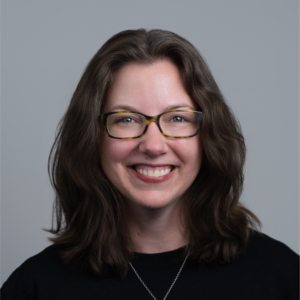 Smiling woman with shoulder-length brown hair, glasses, and a black top against a gray background.