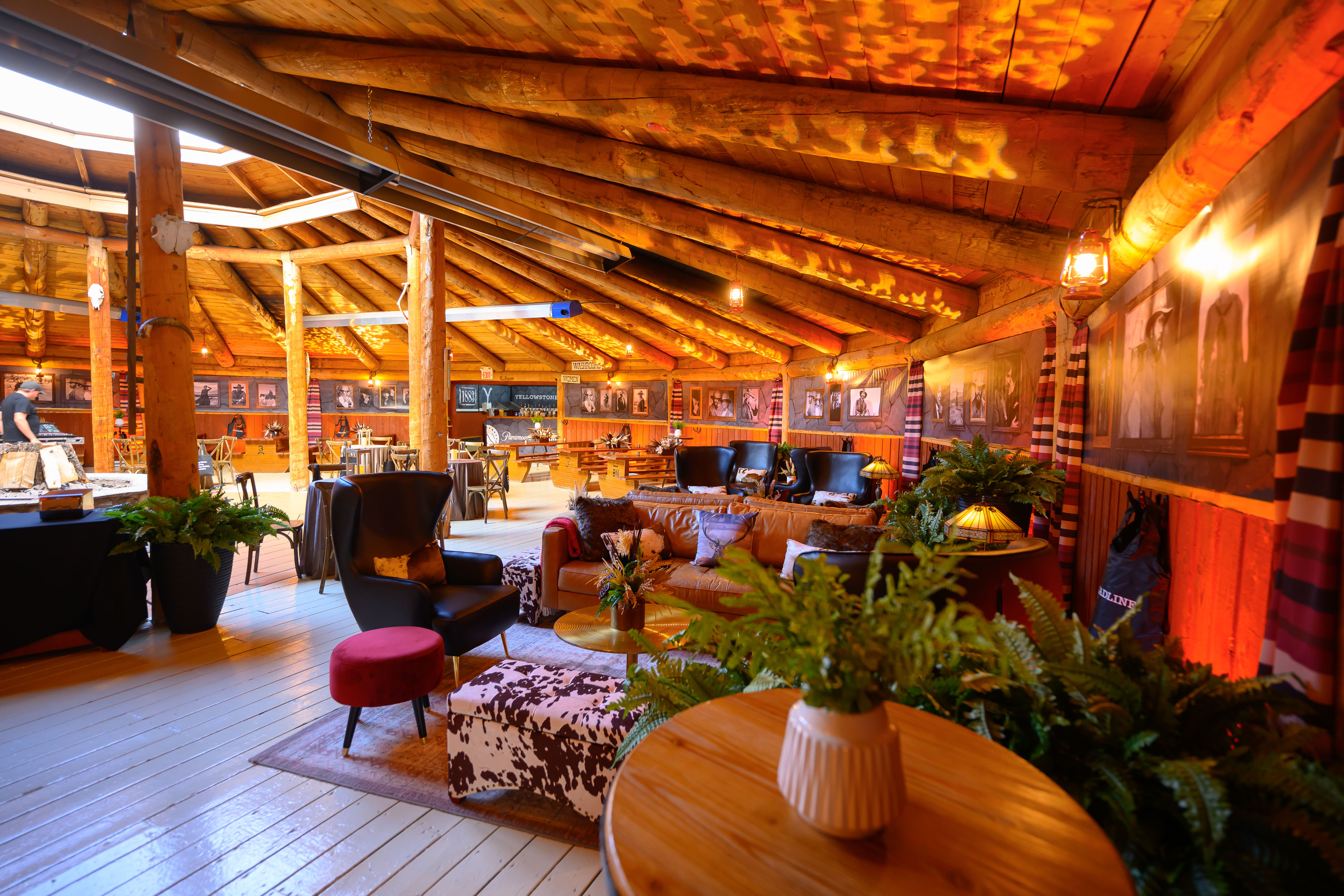 Cozy rustic lodge interior with wooden beams, leather sofas, black armchairs, cowhide ottomans, and warm lighting.