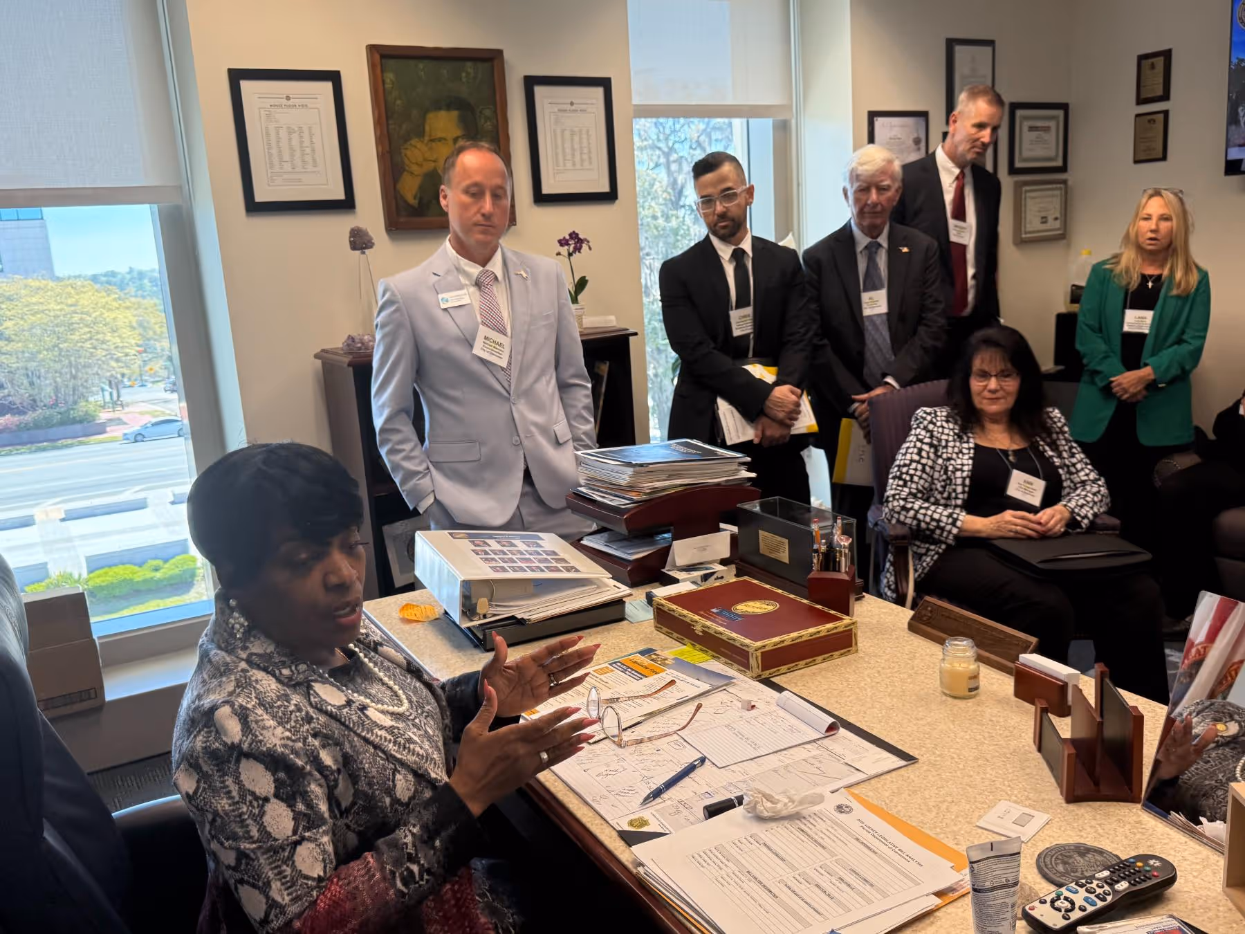 Group of six people in business attire in an office, one woman speaking with animated hand gestures while others listen attentively.
