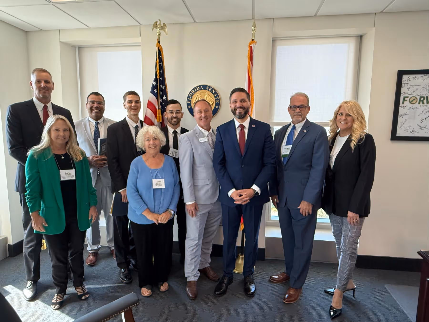 Group of ten professionally dressed people standing in an office with Florida Senate seal and flags in the background.