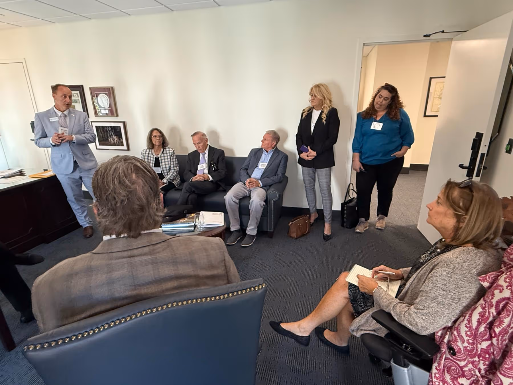 Group of seven adults in a small office room, with one man standing and speaking while others listen seated or standing.